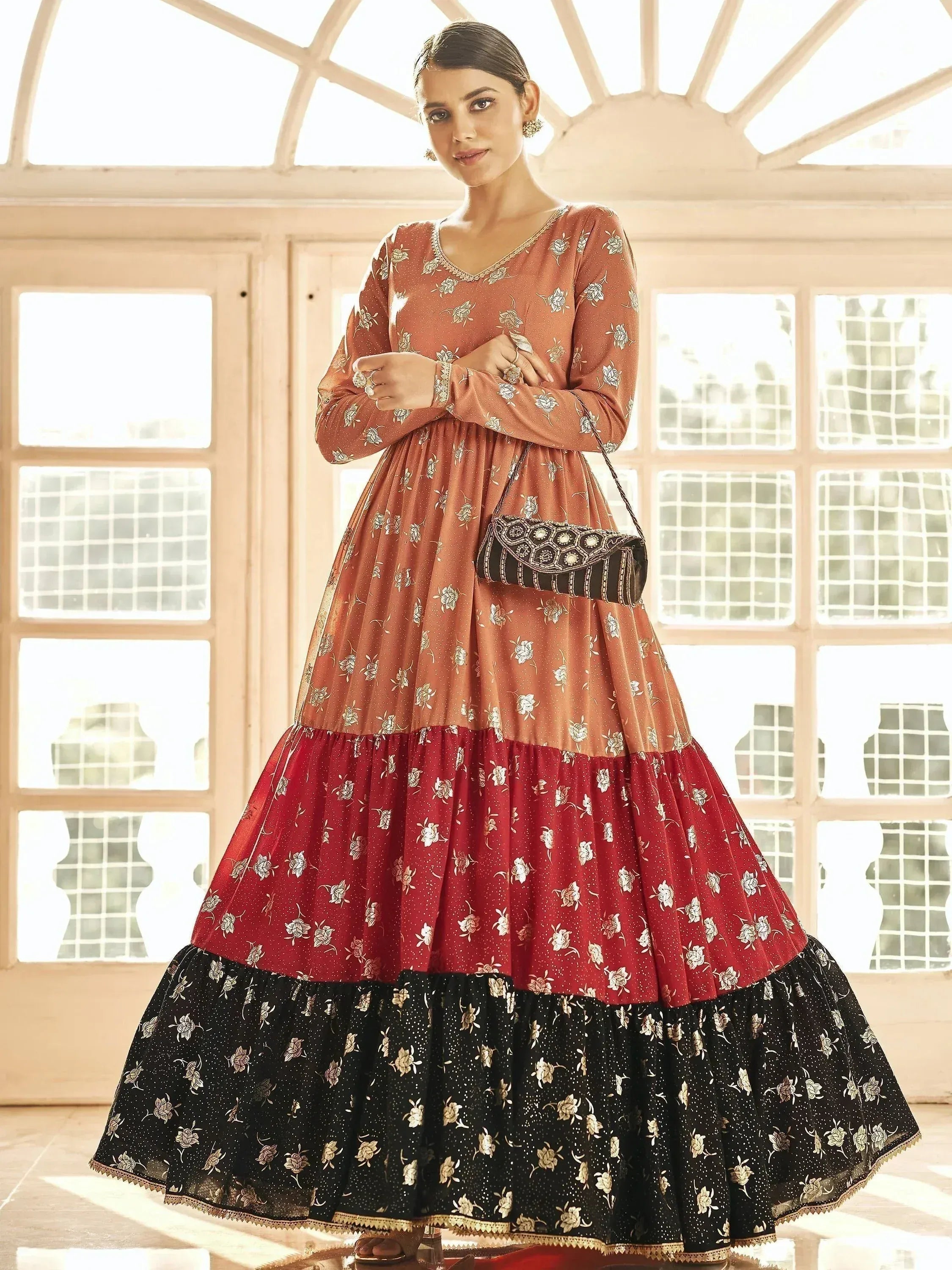 Woman in elegant Indian ethnic gown with floral print, tiered skirt in rust, red, and black, perfect for festive occasions.