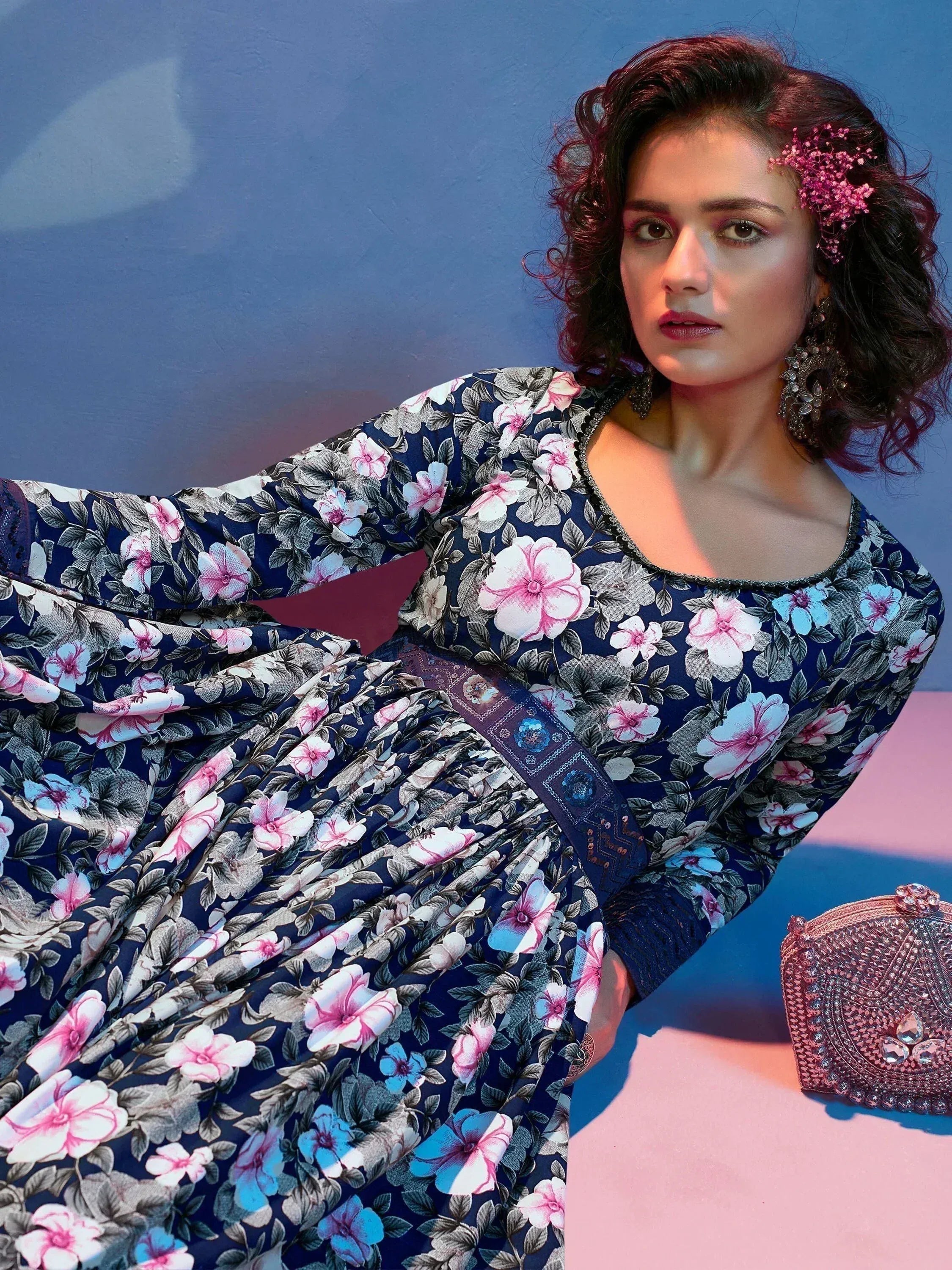 Woman in navy blue floral ethnic gown with pink and white flowers, styled with earrings and a beaded clutch.