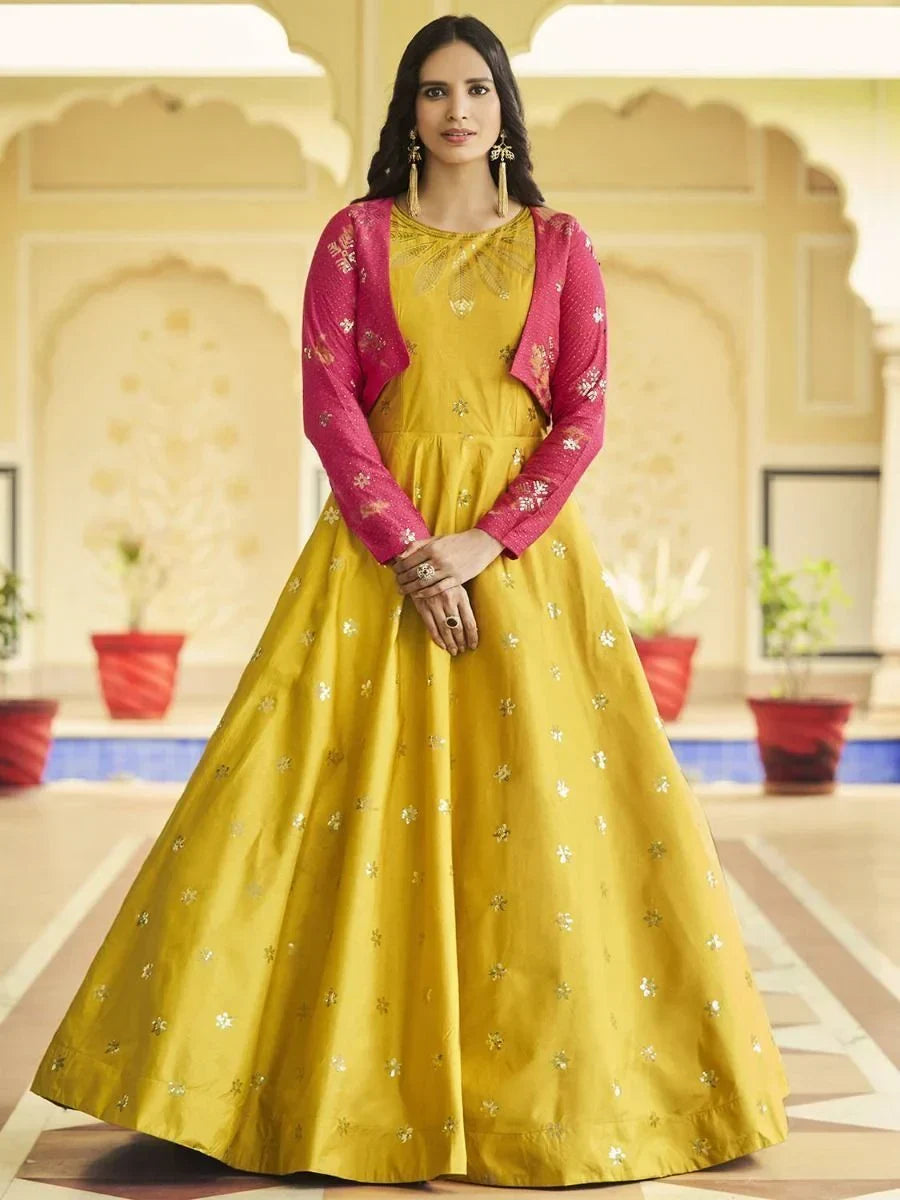 Woman in yellow embroidered ethnic gown with pink jacket, standing in a traditional Indian courtyard