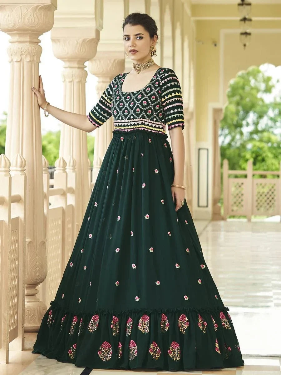 Woman in an embroidered dark green ethnic gown with floral patterns, standing in a corridor.