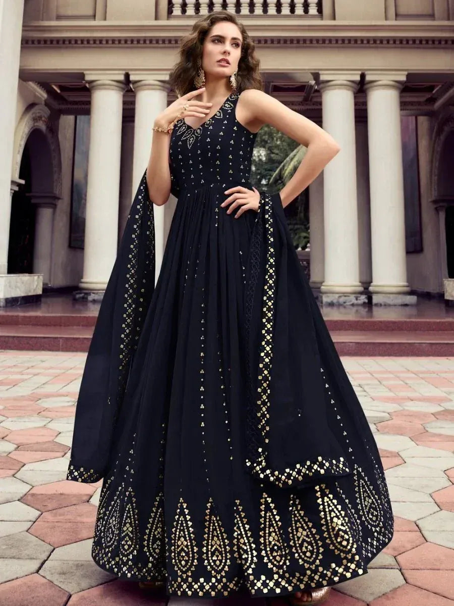 Woman wearing a black ethnic gown with gold embroidery, standing outdoors by pillars.