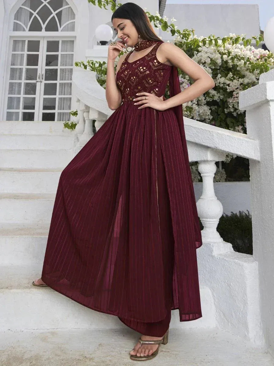 Woman wearing maroon designer ethnic gown with gold detailing, standing outdoors near white stairs.