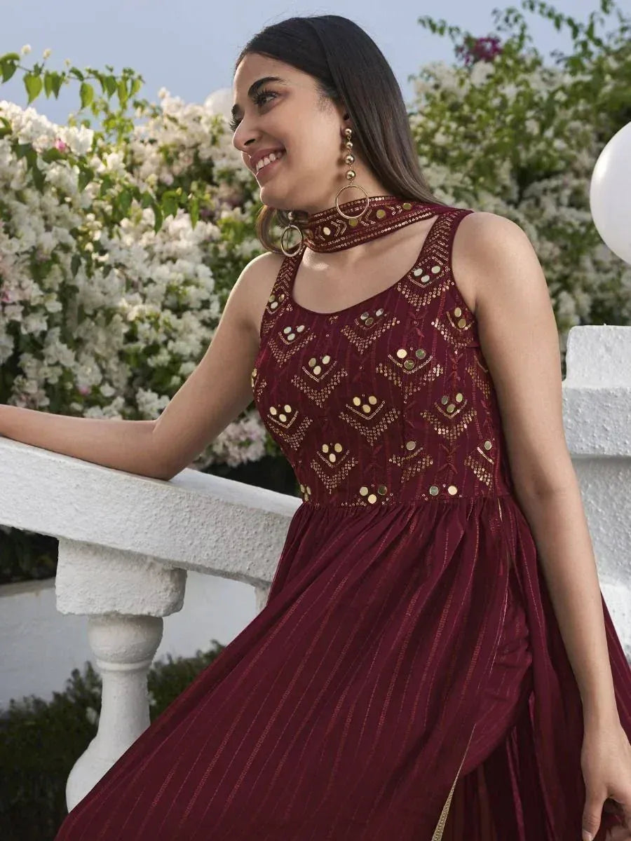 Woman wearing maroon embroidered ethnic gown with gold mirror work, outdoor floral background