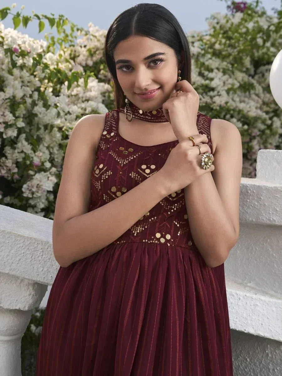 Woman in maroon sleeveless ethnic gown with gold mirror work, posing outdoors with floral background