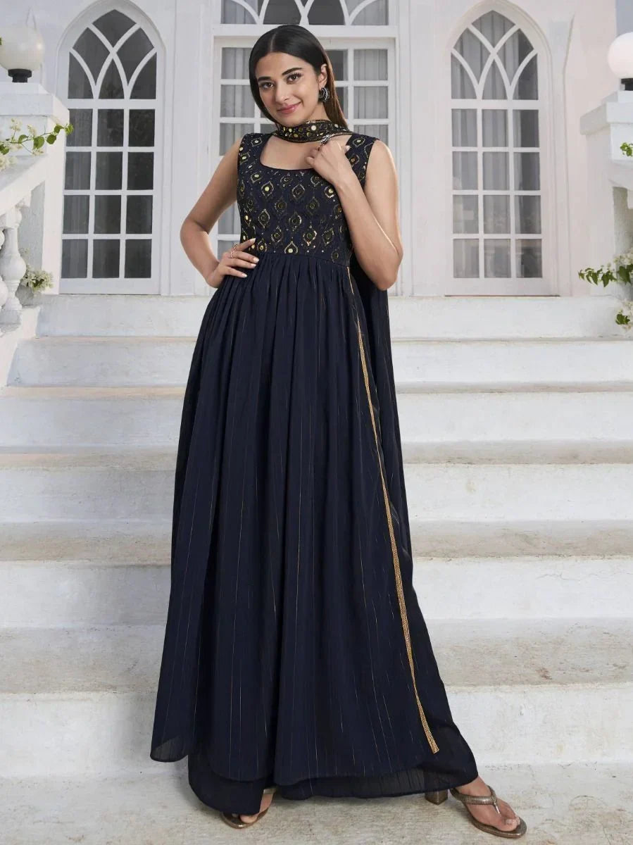 Woman in sleeveless navy blue ethnic gown with gold embroidery, standing on white stairs