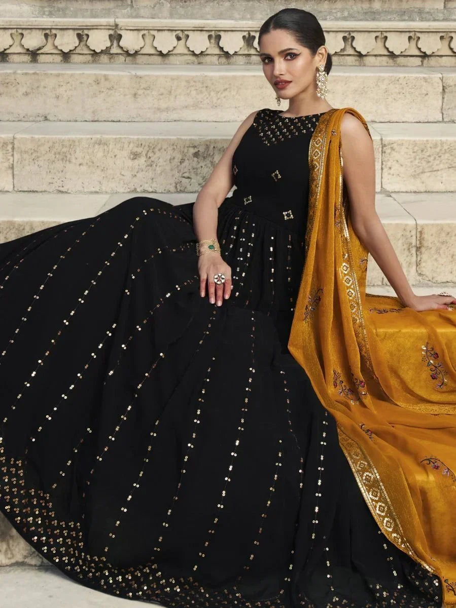 Woman wearing black designer lehenga choli with gold sequin work and mustard dupatta, seated on stone steps.