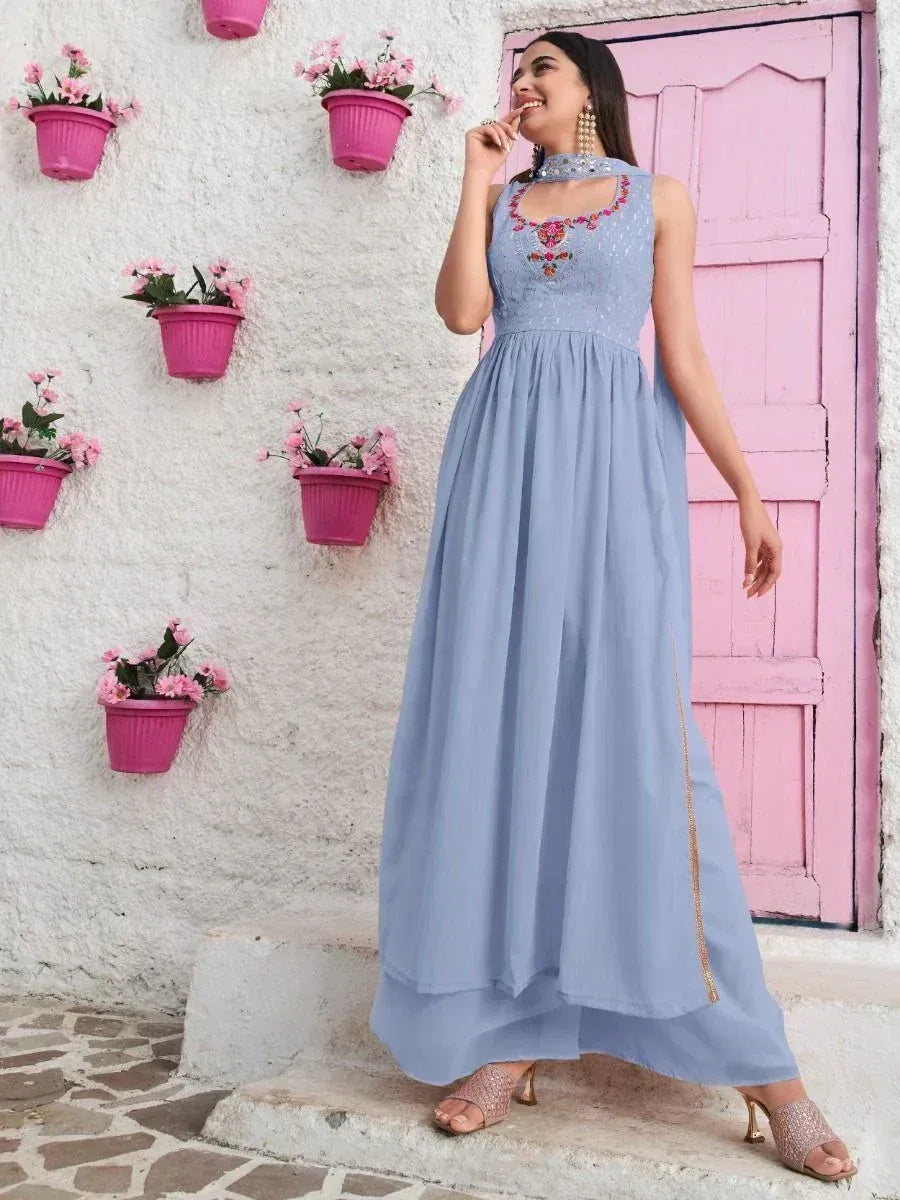 Woman in a light blue ethnic gown with floral embroidery, standing by a pink door and white wall decorated with pink flower pots