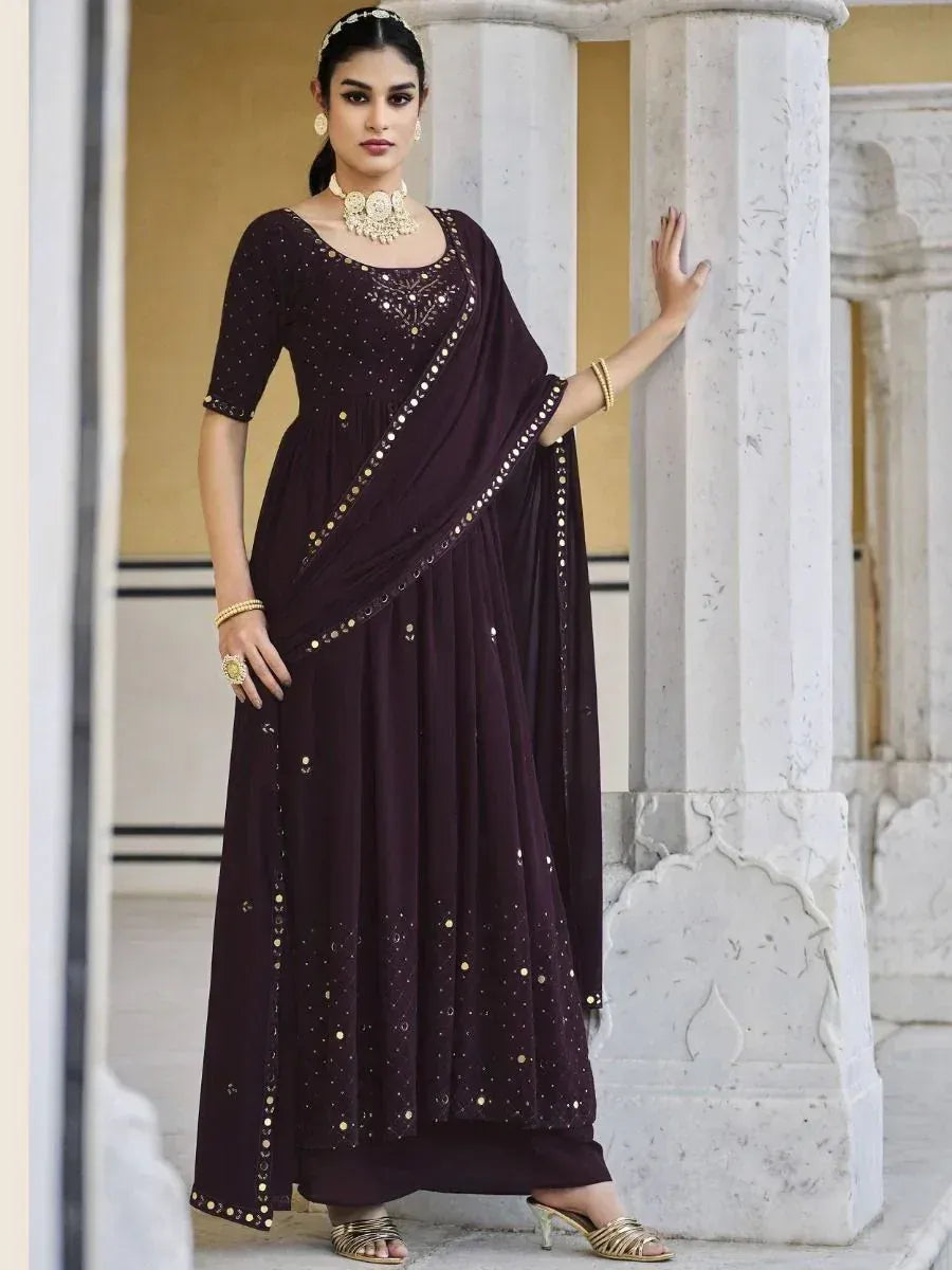 Woman wearing a dark maroon embroidered Anarkali suit with gold mirror work, styled with ethnic jewelry, standing by marble columns
