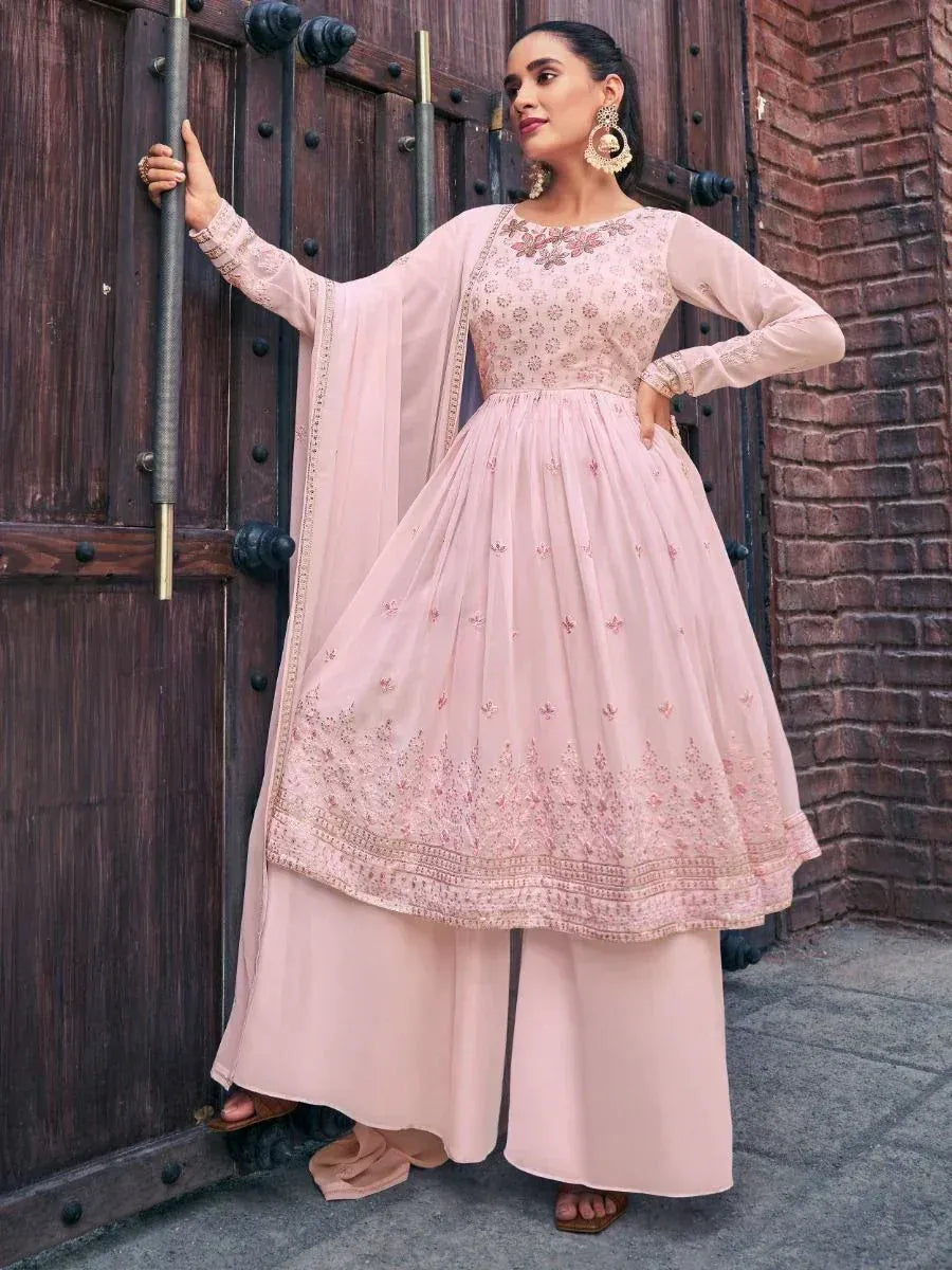 Woman in pastel pink embroidered Anarkali suit with dupatta, standing by wooden door