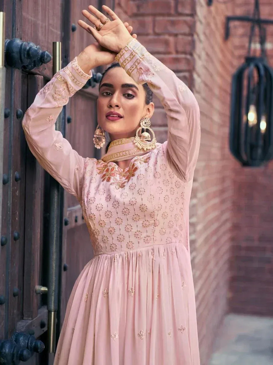 Woman in pastel pink embroidered Anarkali gown with long sleeves, statement earrings, standing near a wooden door