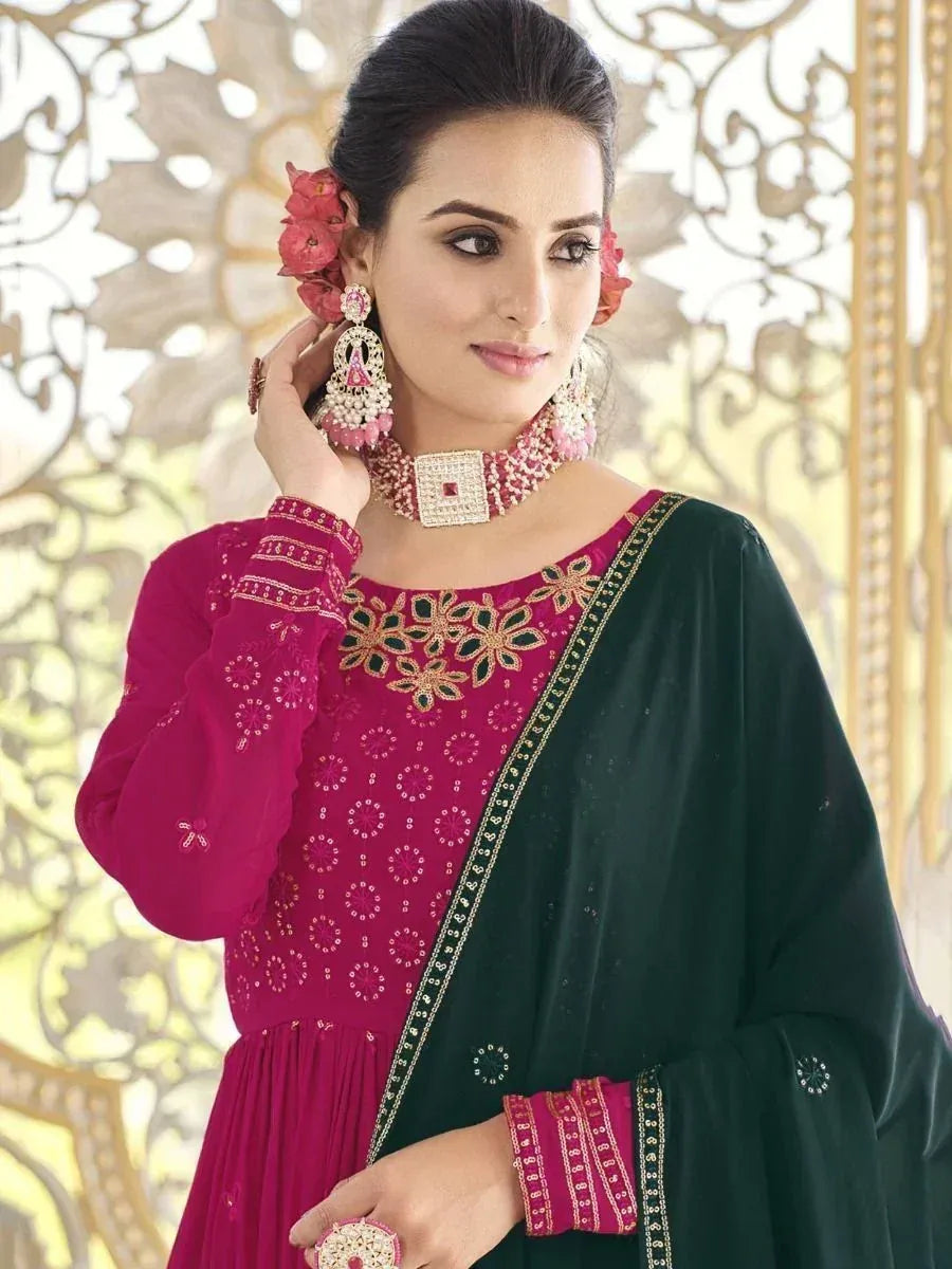 Woman in embroidered magenta and green ethnic gown with floral jewelry, styled for Indian occasion.