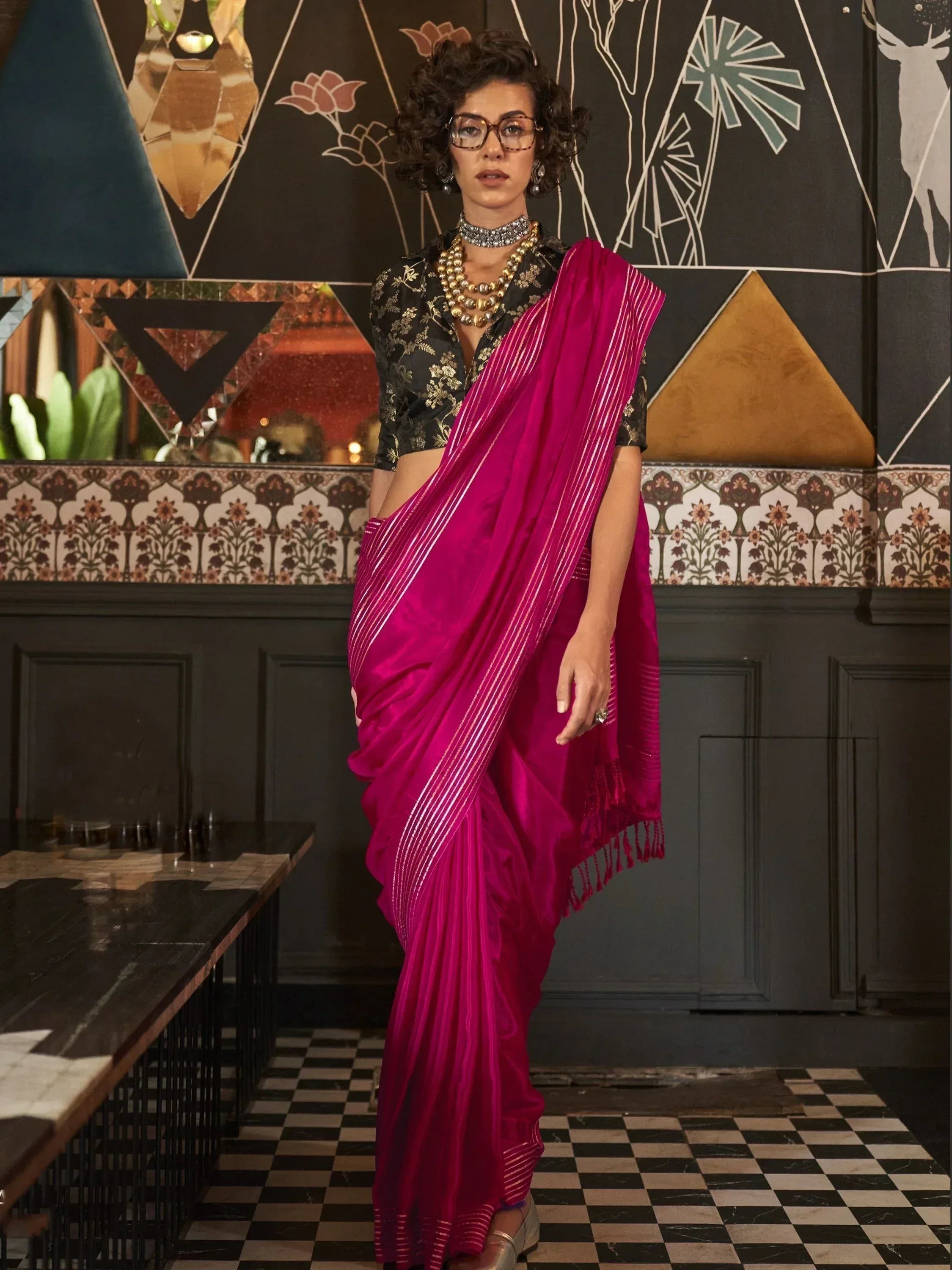Vibrant pink saree with gold stripes, paired with black floral blouse and statement jewelry.