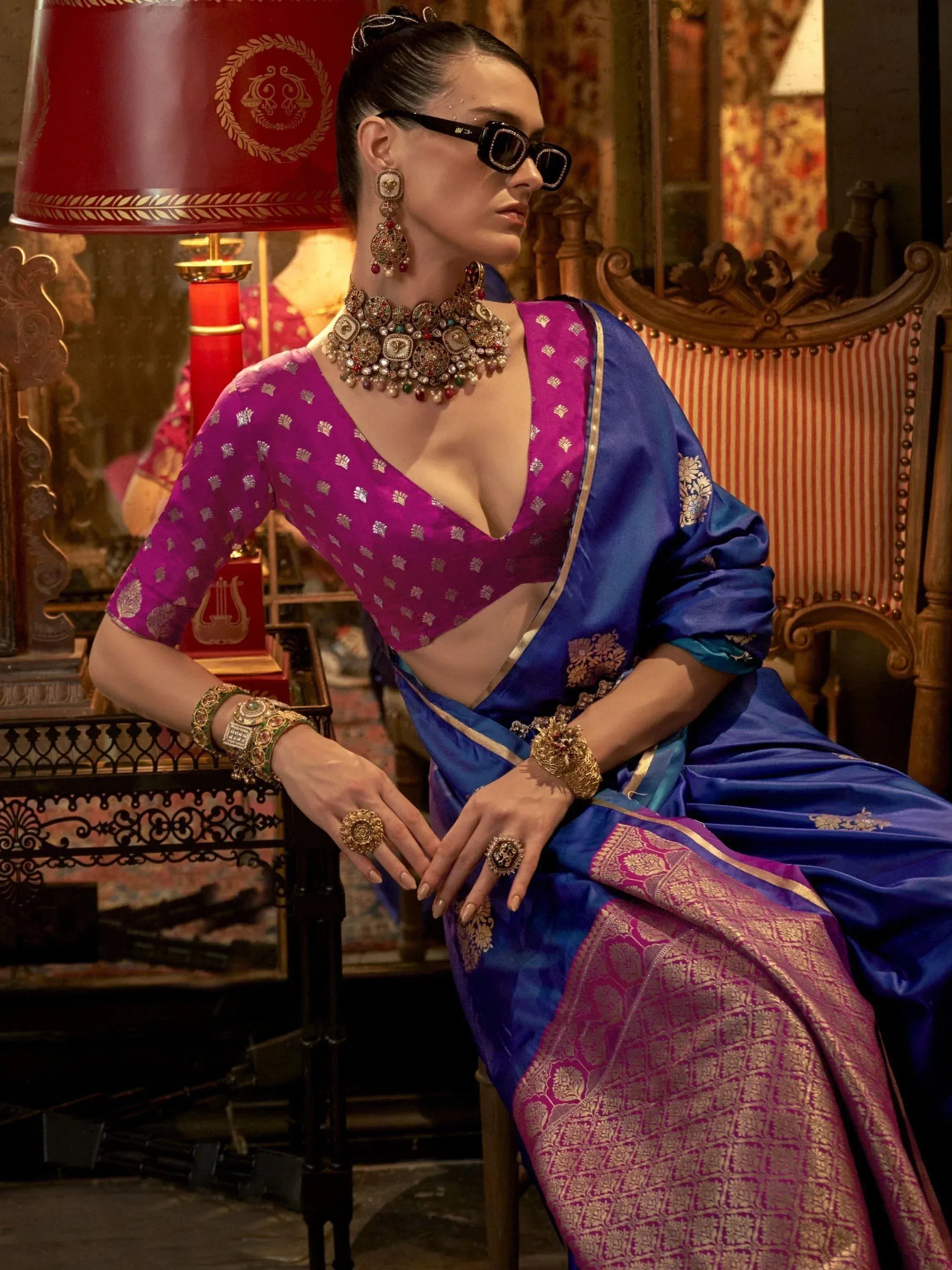 Elegant woman in royal blue and magenta designer saree with gold embroidery, statement jewelry, and sunglasses.