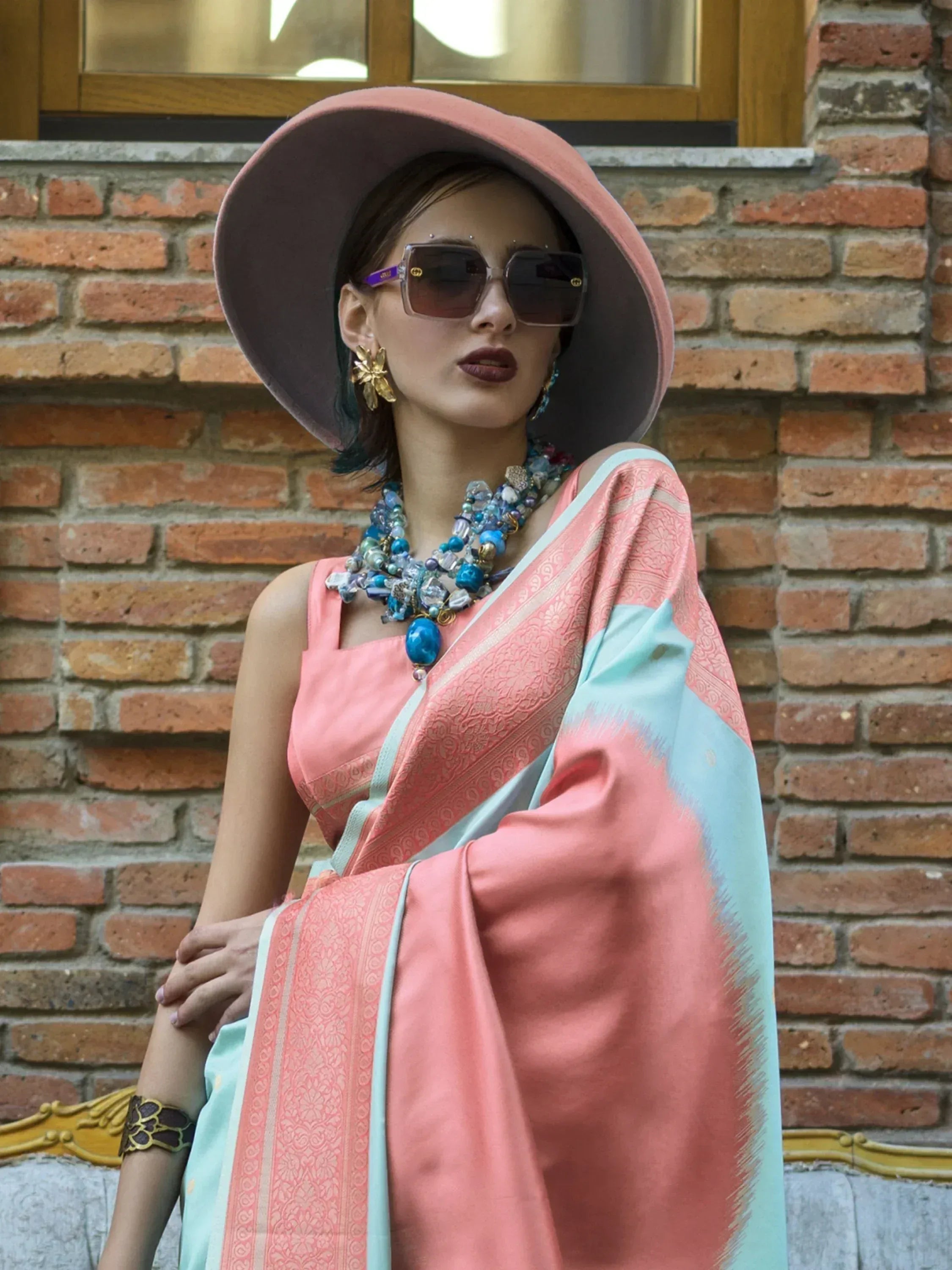 Woman in pink and mint saree with wide-brim hat, statement jewelry, and sunglasses against brick wall