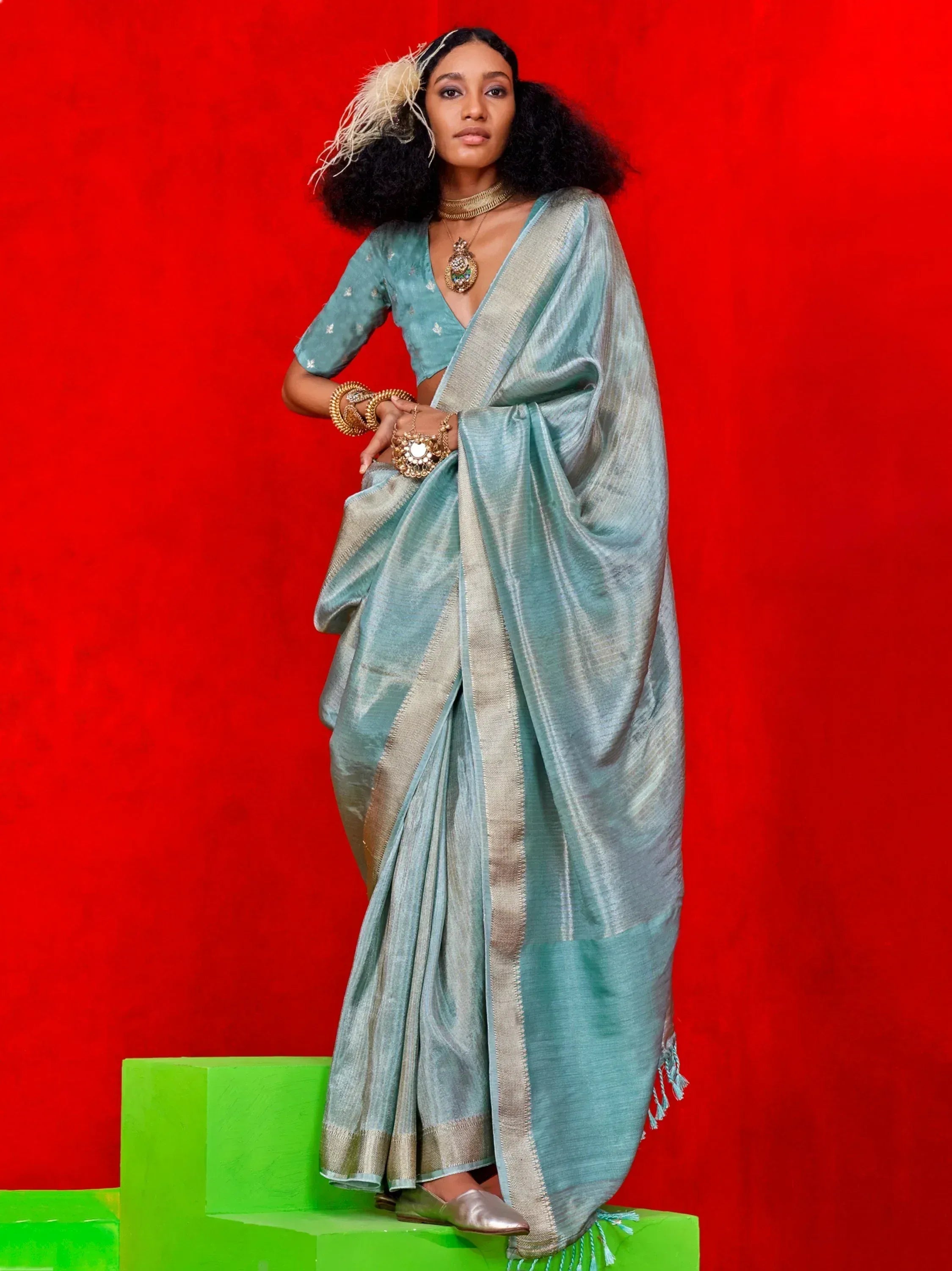 Woman in elegant light blue saree with gold border, styled with traditional jewelry against red backdrop