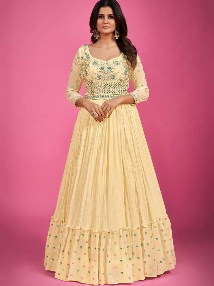 Woman in a yellow embroidered Anarkali gown with floral patterns, standing against a pink background