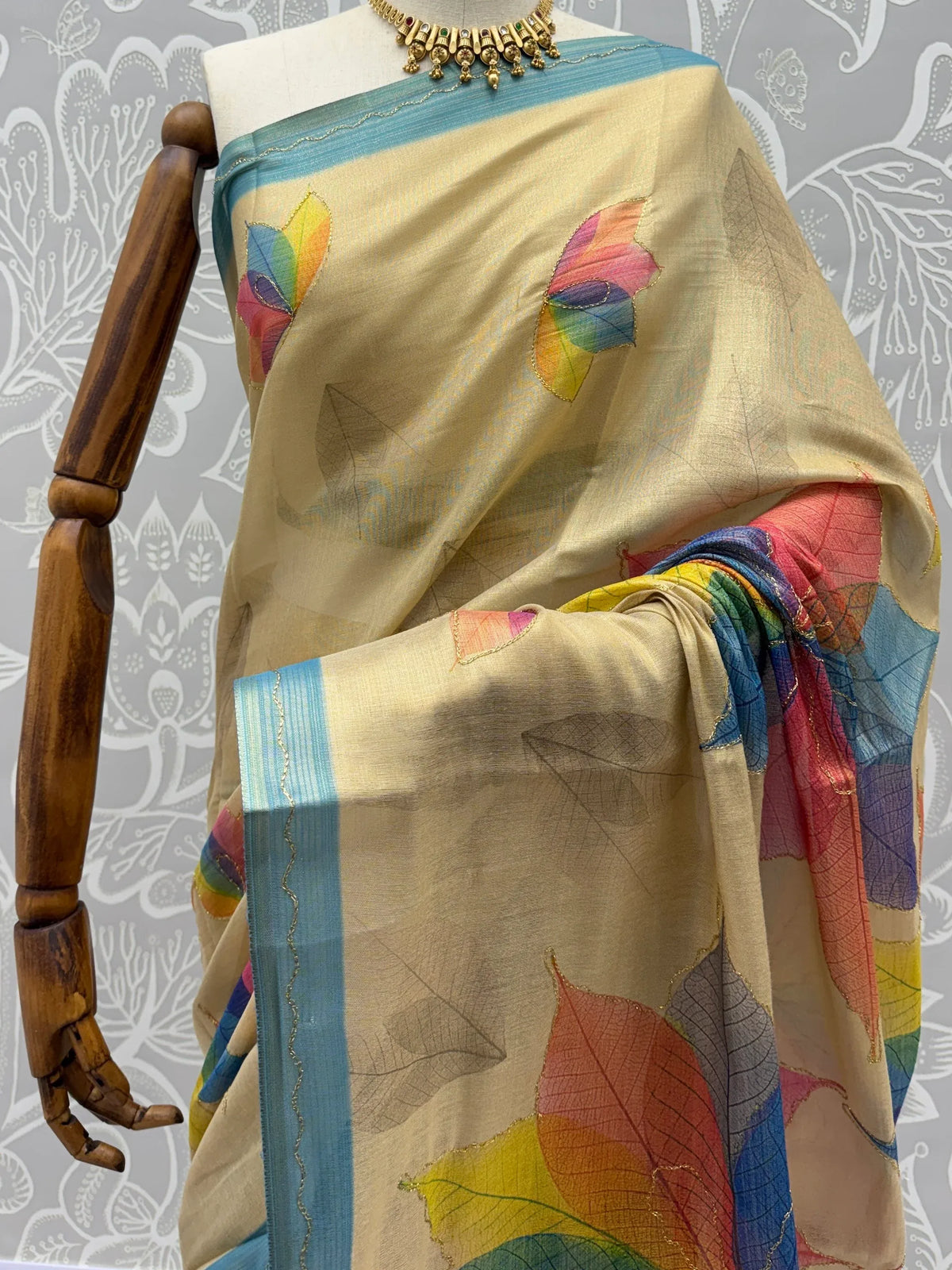 Beige designer saree with colorful leaf motifs and a blue border on a mannequin