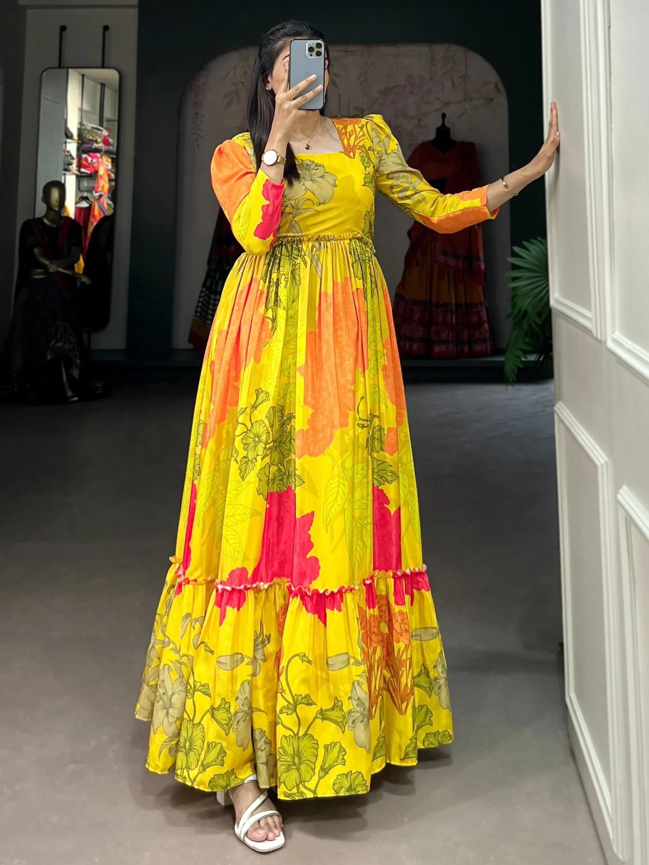 Woman in a yellow floral printed ethnic gown with long sleeves and tiered hem, standing indoors