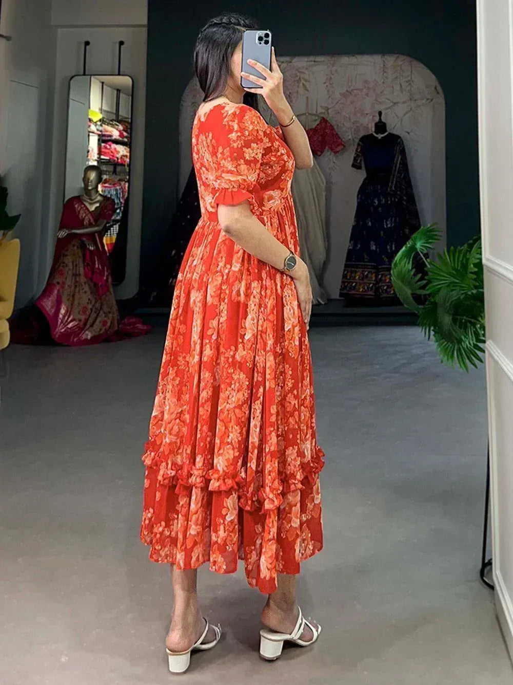 Woman in orange floral ethnic gown with ruffled hem, taking mirror selfie in boutique