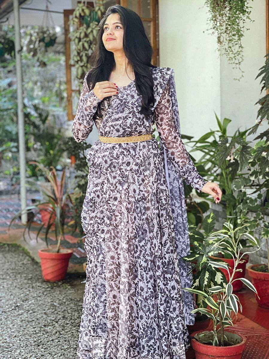 Woman in floral ethnic gown with sheer sleeves and gold belt, surrounded by potted plants