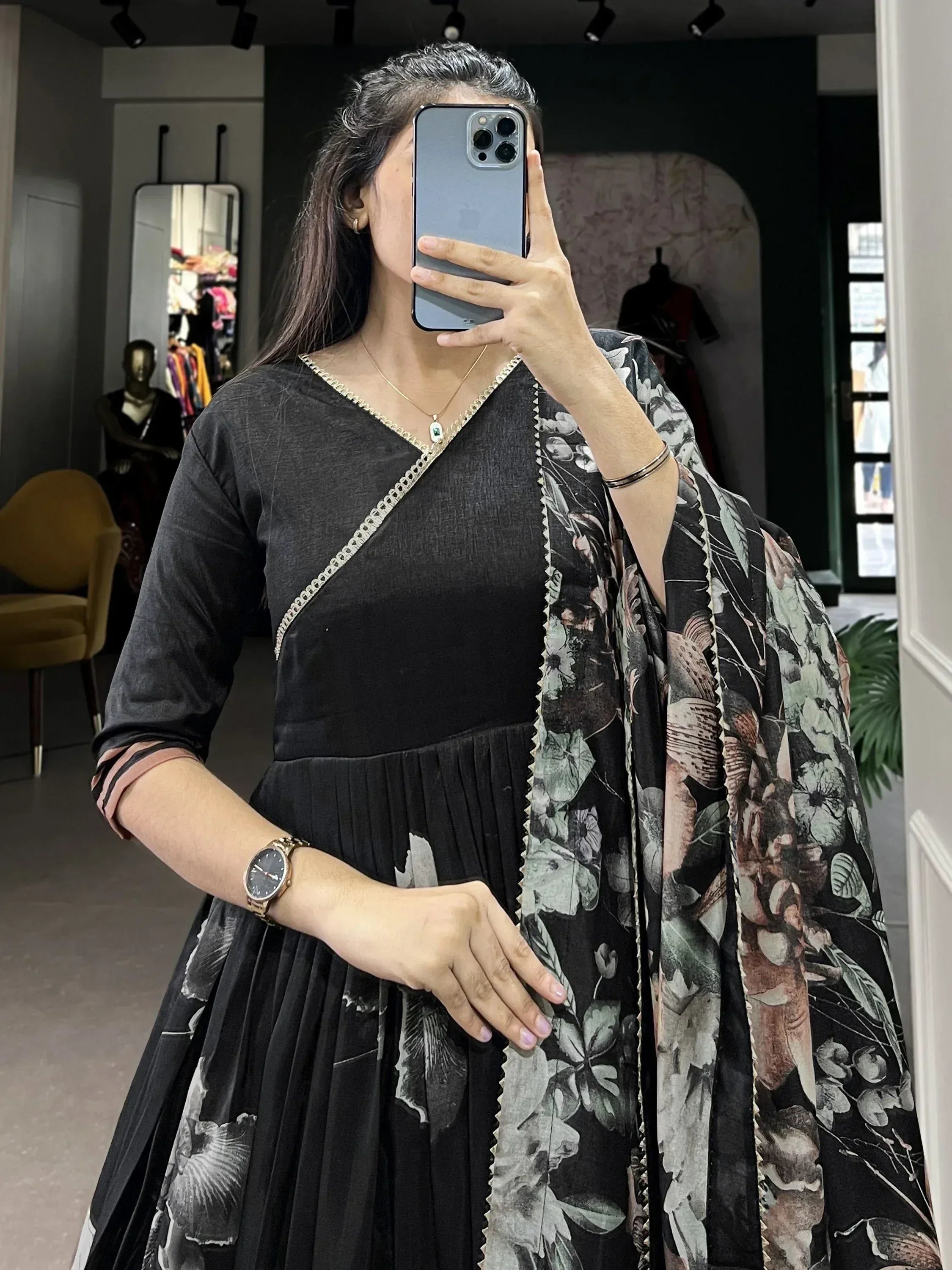 Woman in black floral print ethnic gown with gold trim, taking mirror selfie in boutique