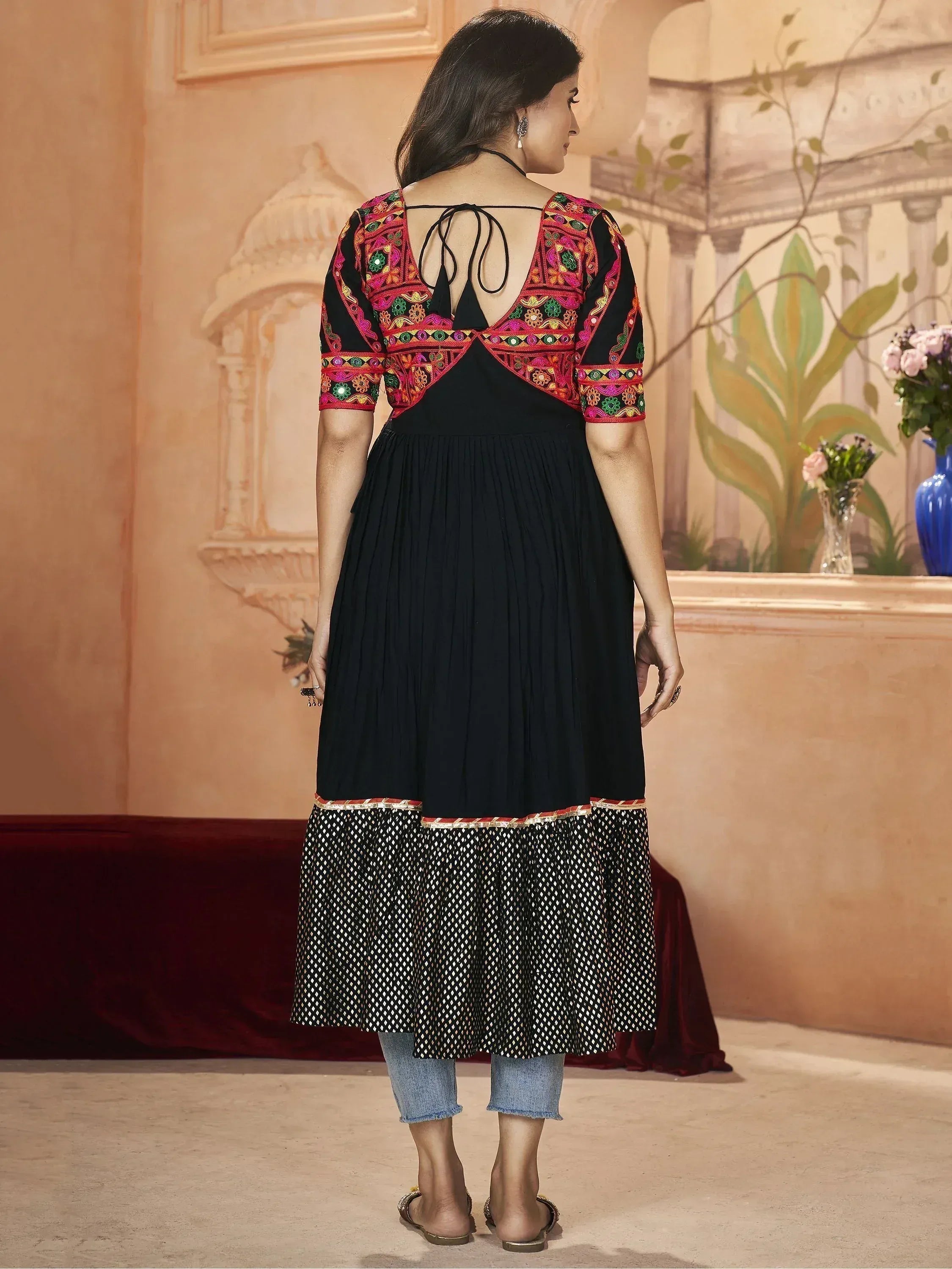 Black ethnic gown with colorful embroidery, back tie, and pleated hem, styled with jeans for modern Indian wear.