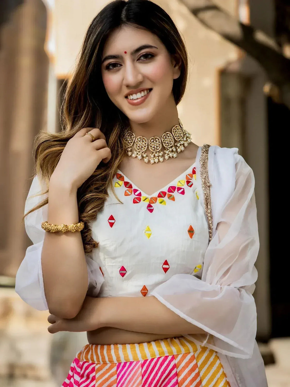 Woman in white embroidered top with colorful geometric patterns and striped skirt, accessorized with gold jewelry