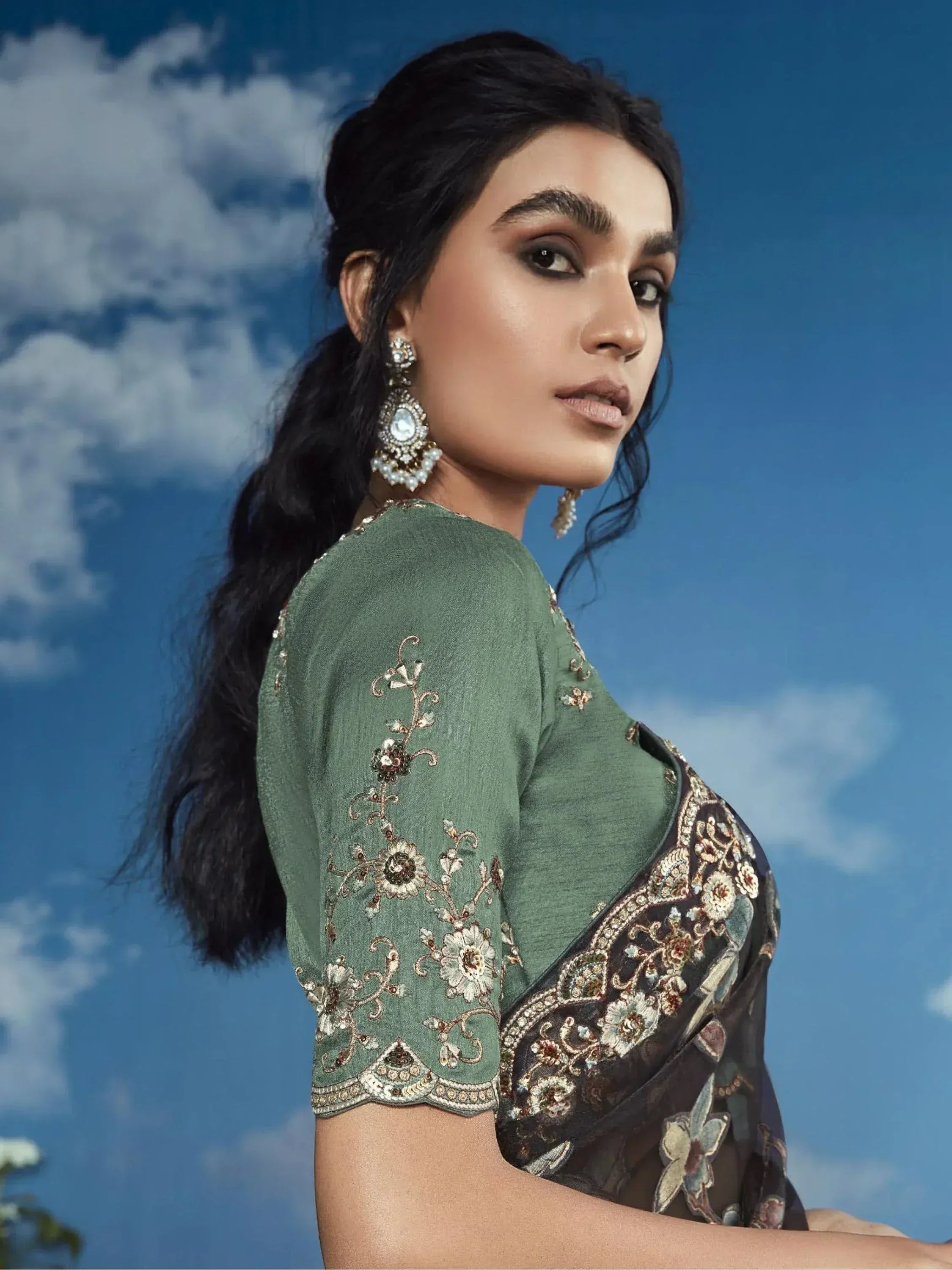 Woman in green embroidered blouse with floral design, paired with embellished saree, against blue sky backdrop