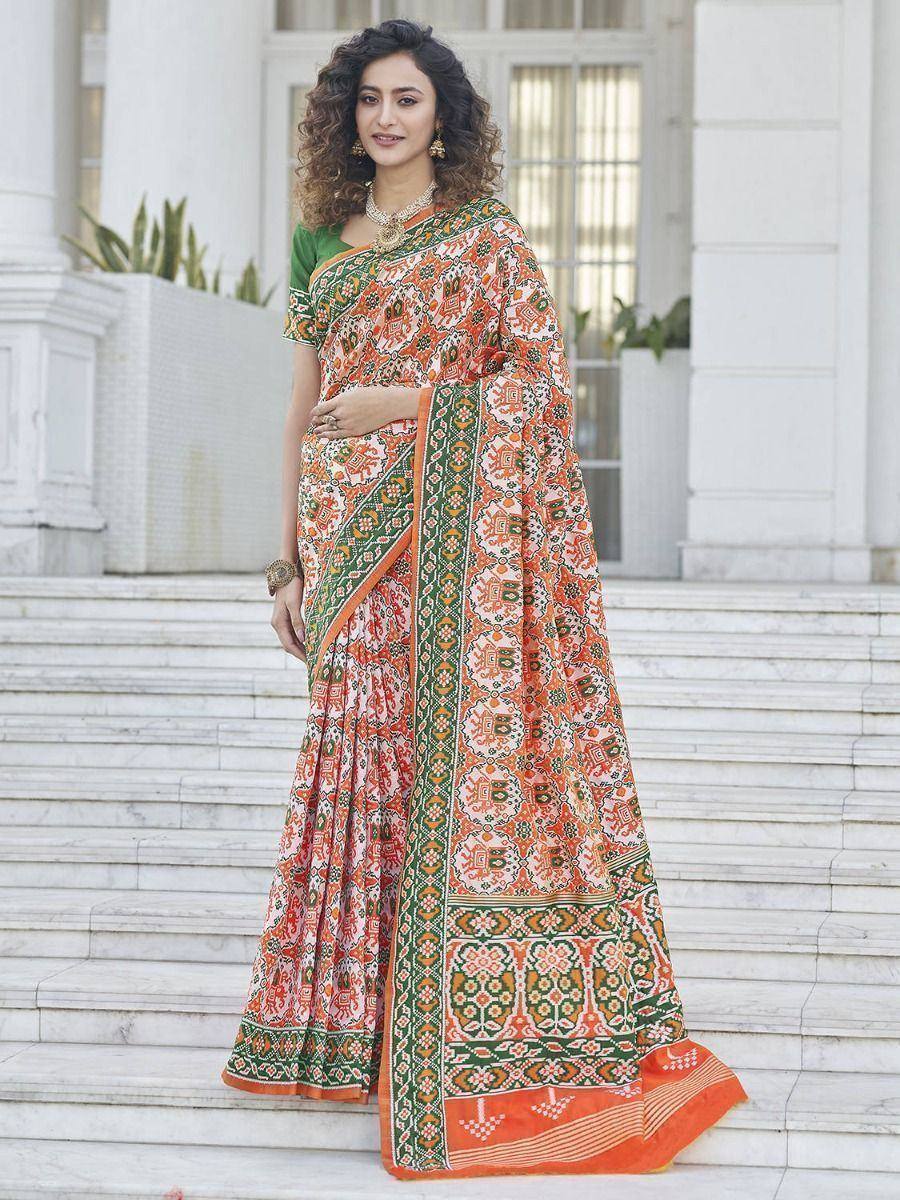 Woman in orange and green printed saree with ethnic motifs, styled with gold jewelry on marble steps