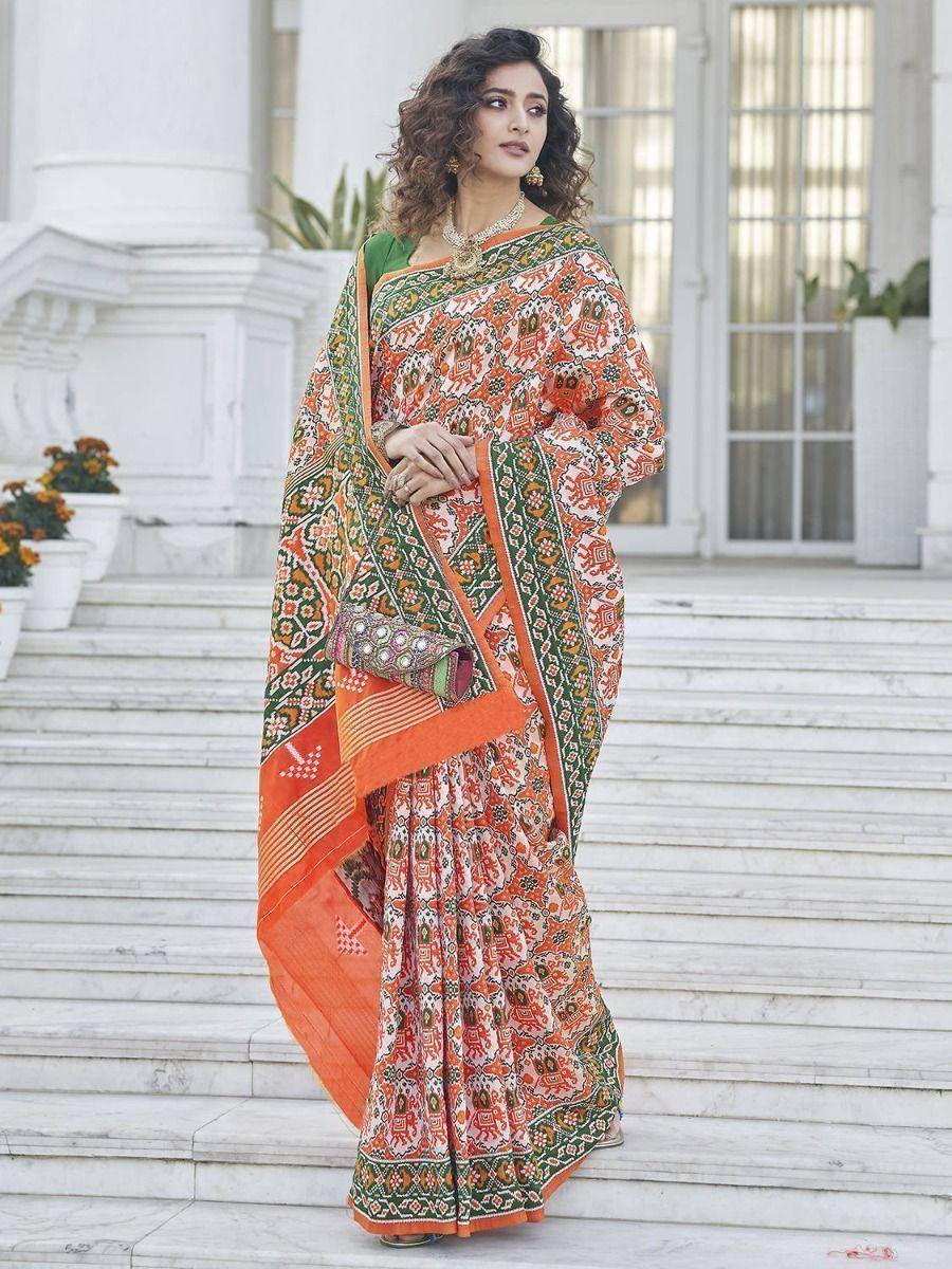 Woman in vibrant orange and green printed saree with ethnic patterns, standing on marble steps
