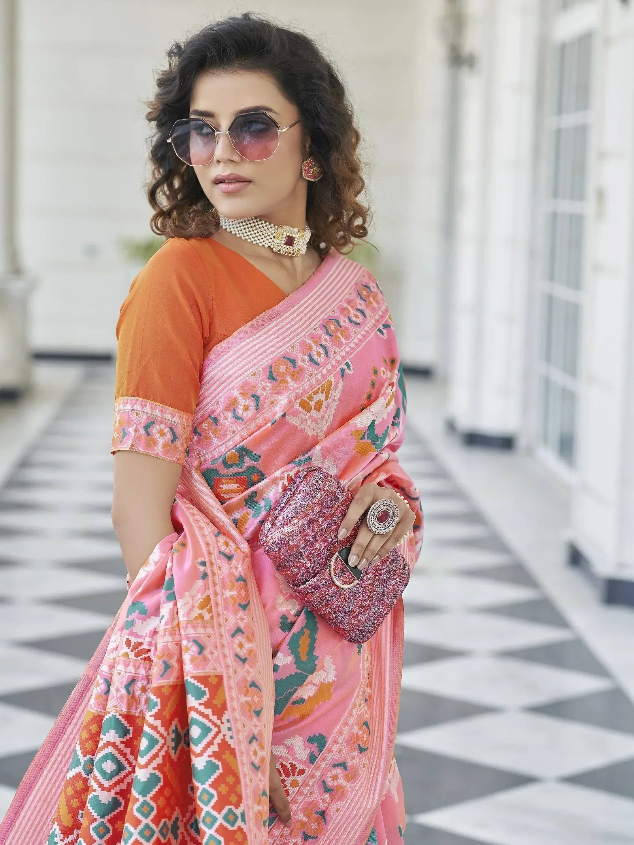 Woman in pink printed saree with orange blouse, ethnic jewelry, and sunglasses, holding glitter clutch