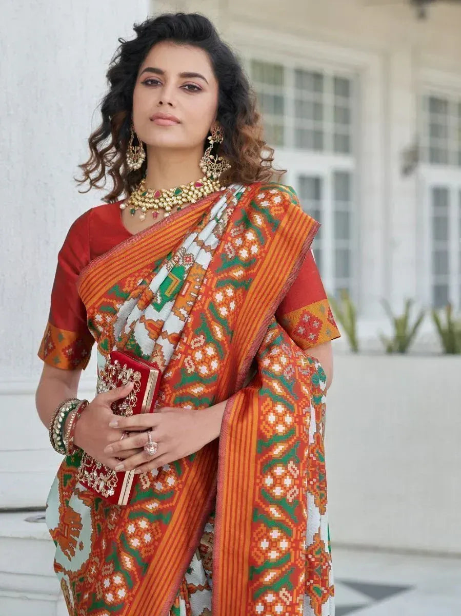 Woman in vibrant orange and green printed saree with red blouse, traditional jewelry, holding clutch