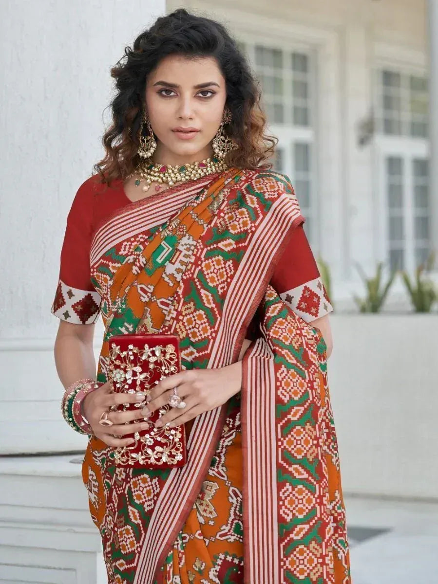 Woman in vibrant red, green, and orange printed saree with matching blouse, holding embellished clutch, styled for ethnic occasions