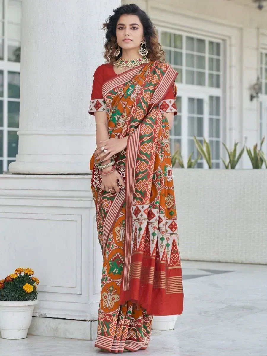 Woman in vibrant printed saree with red blouse, ethnic jewelry, standing by white pillar