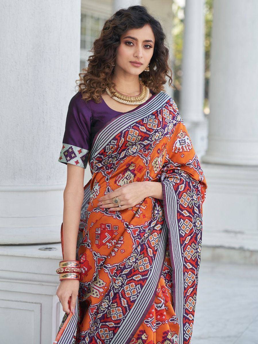 Woman in vibrant orange and purple printed saree with geometric patterns, paired with matching blouse and gold jewelry