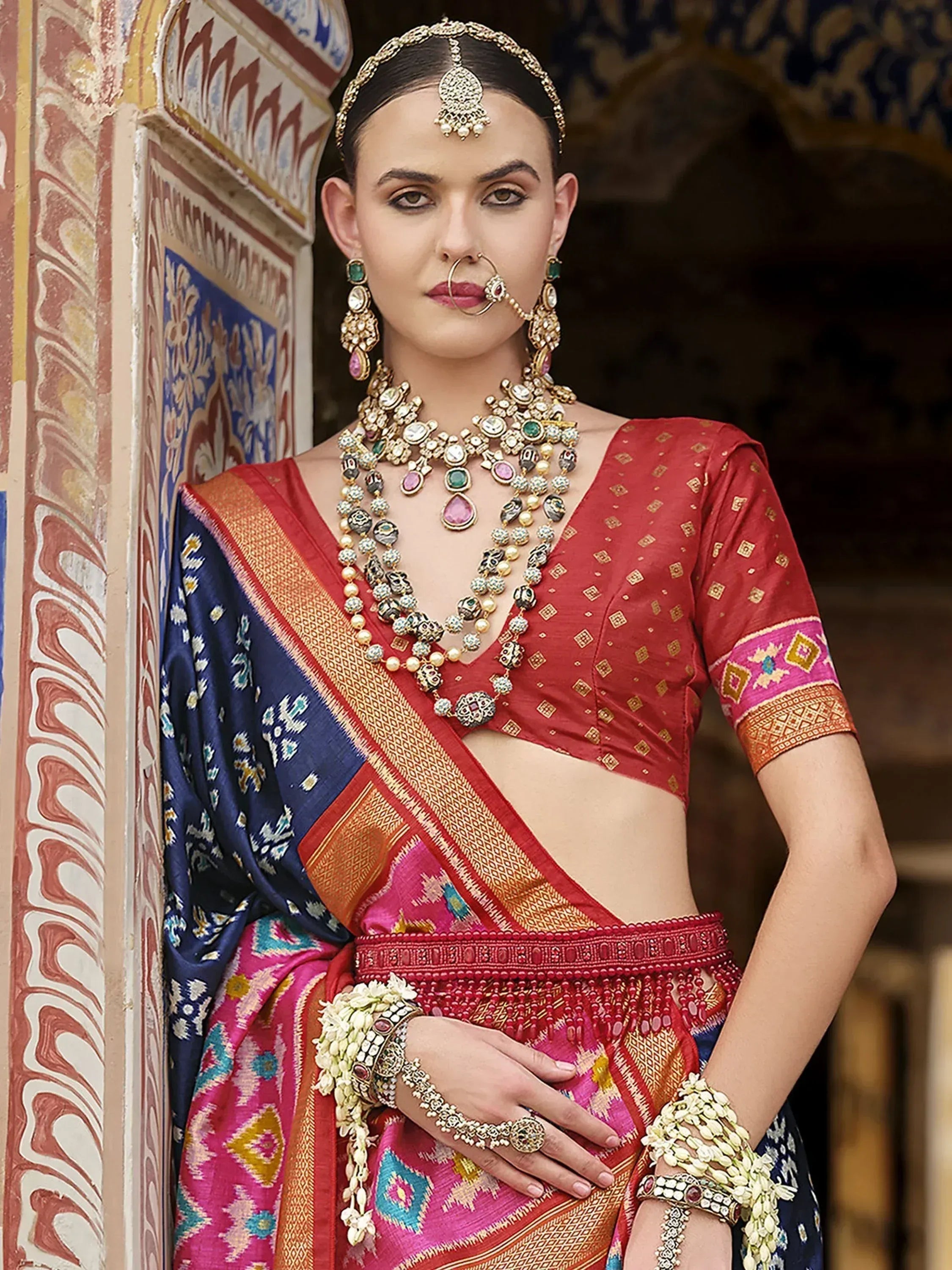 Woman in vibrant red and blue Indian saree with gold embroidery, traditional jewelry, and floral accessories.