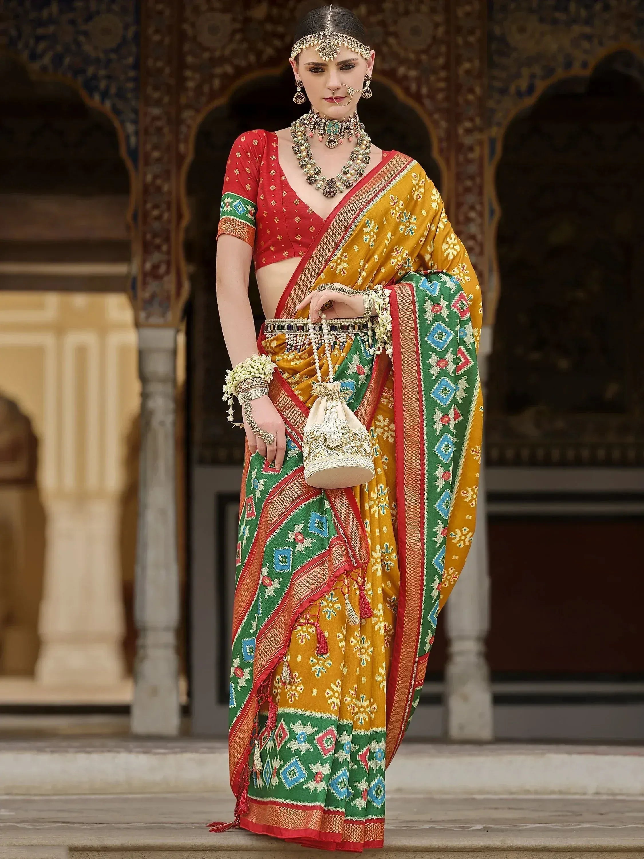 Woman in vibrant yellow and green ethnic saree with red blouse, traditional jewelry, and ornate palace backdrop