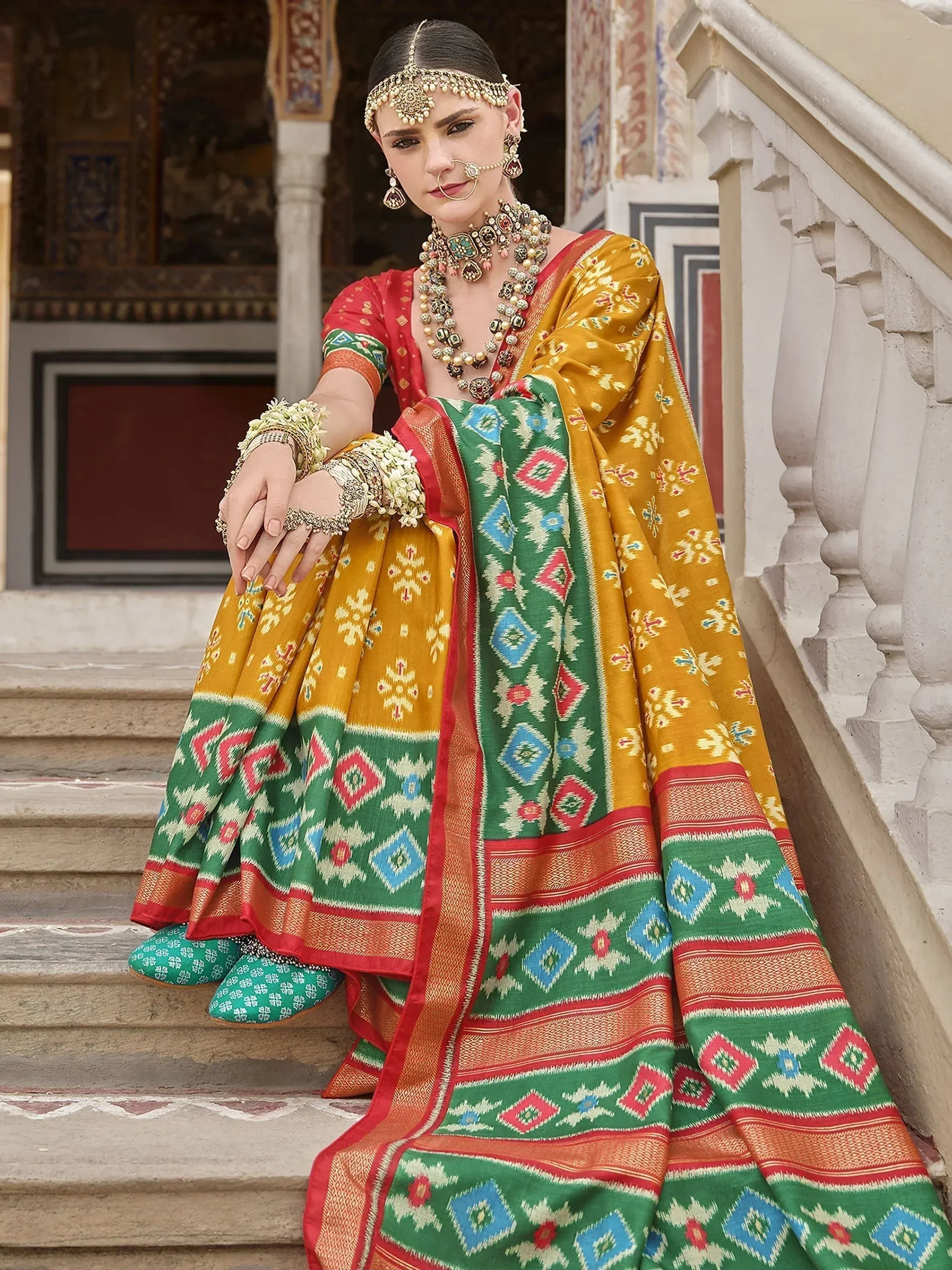 Woman in vibrant yellow and green printed lehenga choli with traditional jewelry, sitting on palace steps