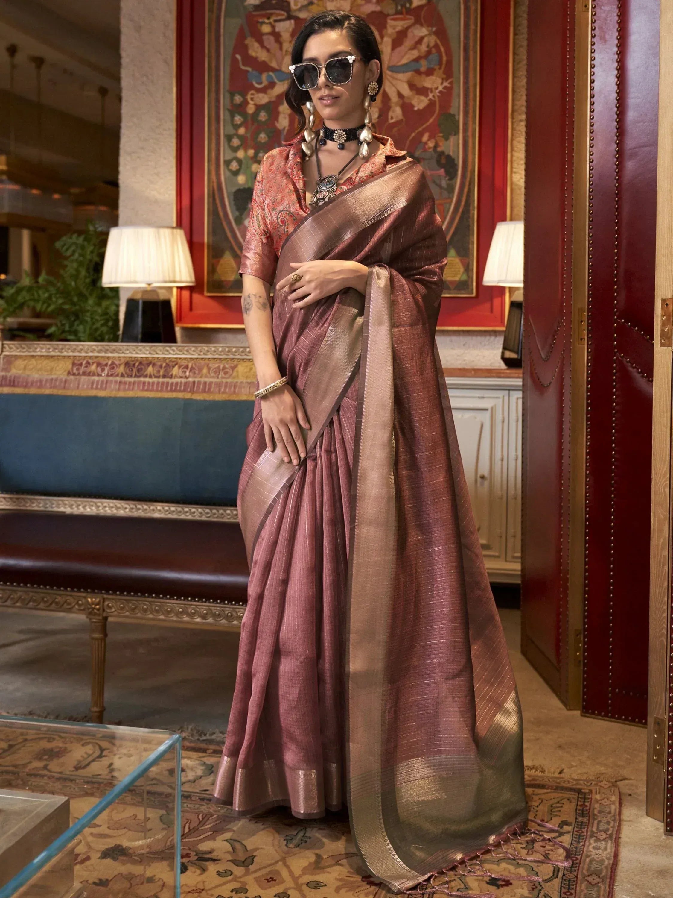Woman in elegant mauve silk saree with gold border, styled with statement jewelry and sunglasses for festive occasions.