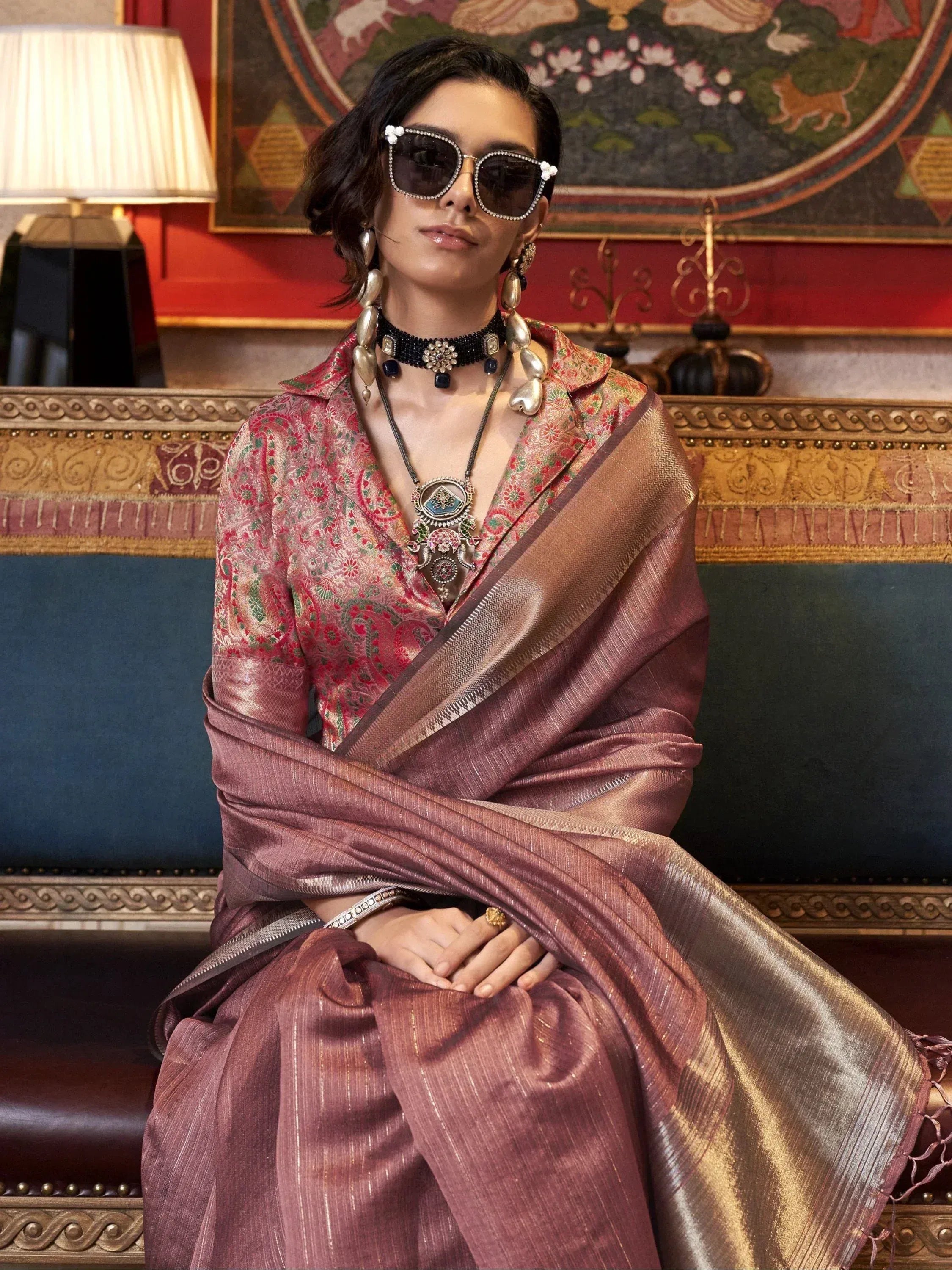 Stylish woman in designer ethnic saree with floral blouse, statement jewelry, and sunglasses, seated elegantly.
