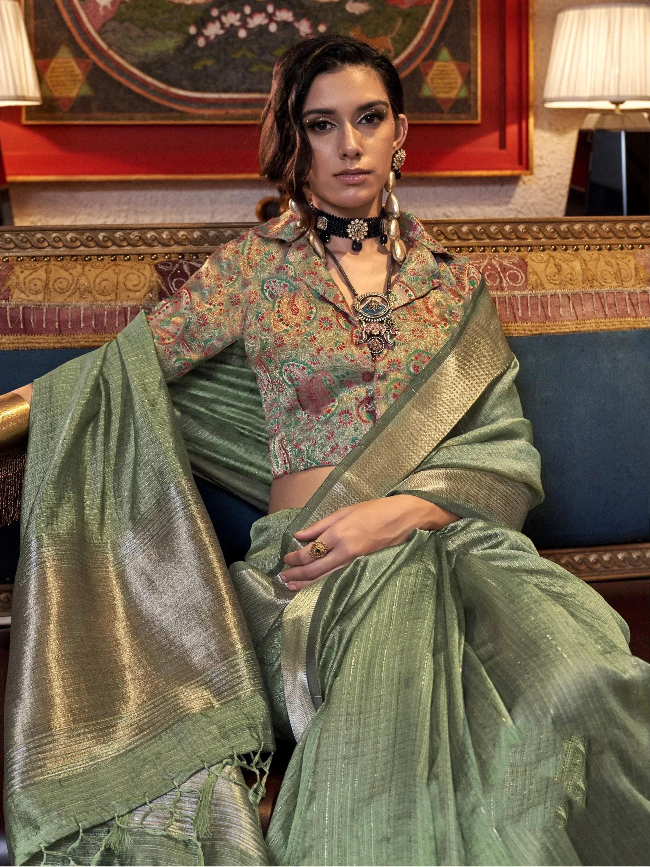 Woman in olive green saree with floral blouse, ethnic jewelry, seated on ornate sofa