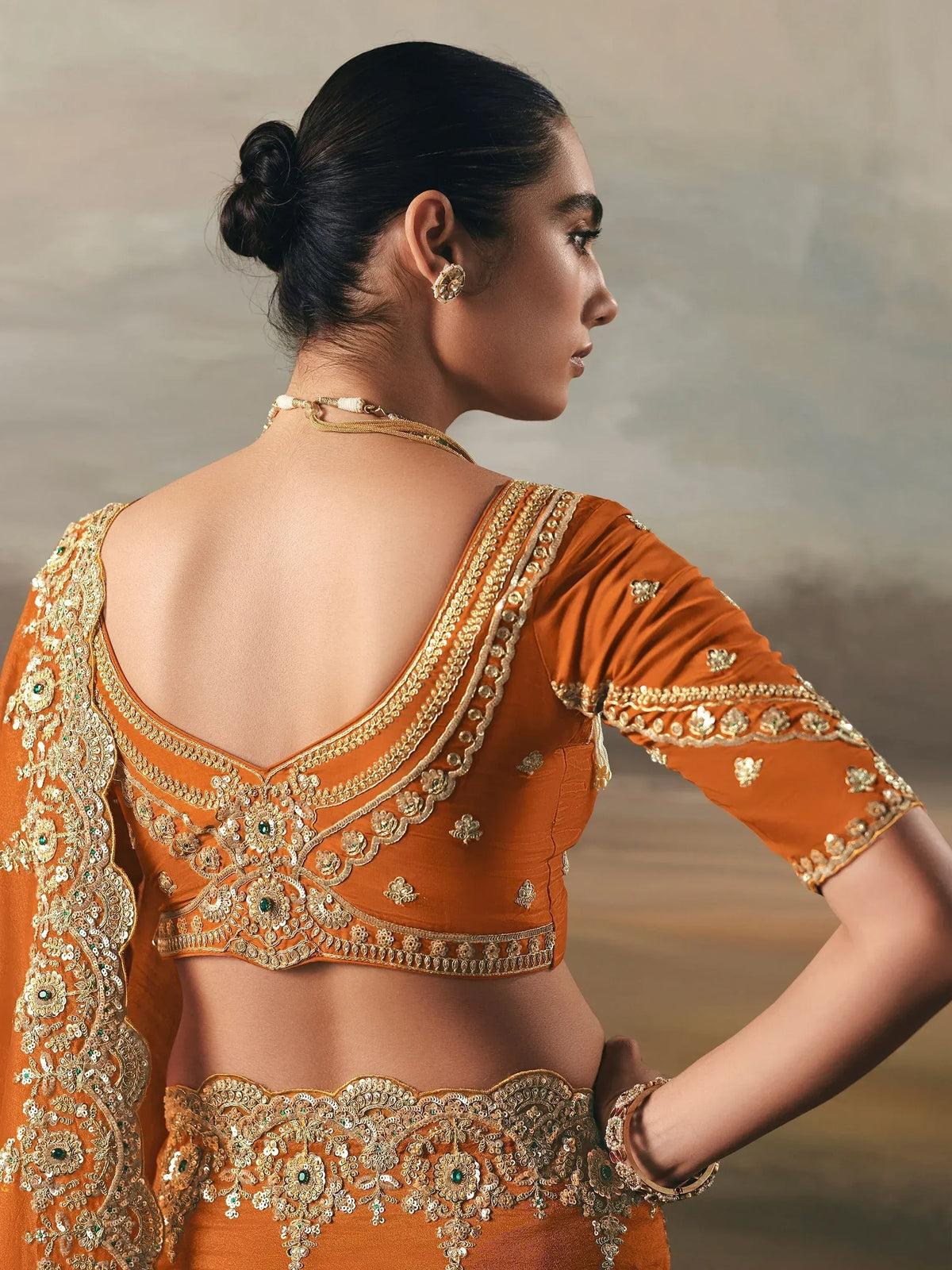 Orange designer lehenga choli with gold embroidery and green accents, showcasing intricate back detailing.