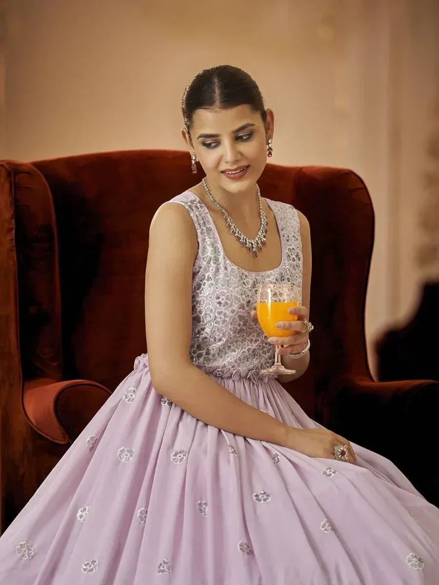 Woman in lavender ethnic gown with floral embroidery, holding orange juice, seated on velvet chair