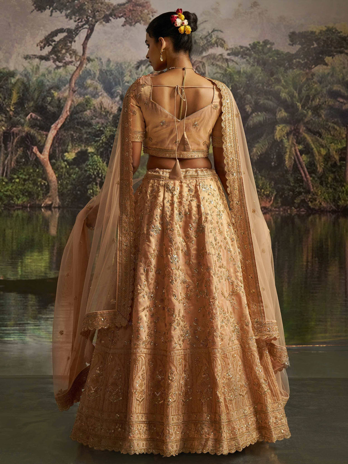 Elegant peach lehenga choli with gold embroidery, sheer dupatta, floral hair accessory, back view near water