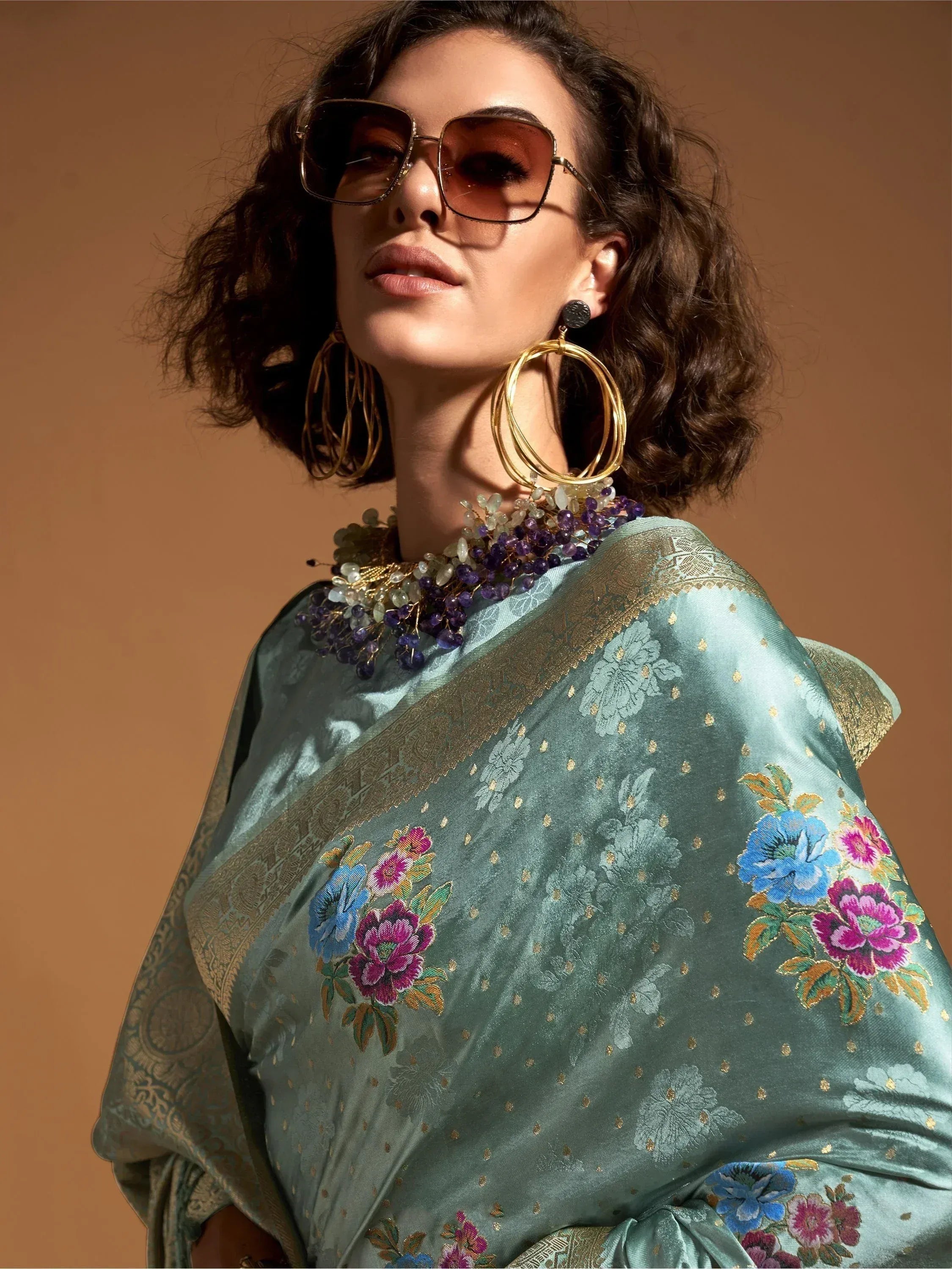 Stylish woman in teal floral embroidered saree with gold border, statement jewelry, and sunglasses