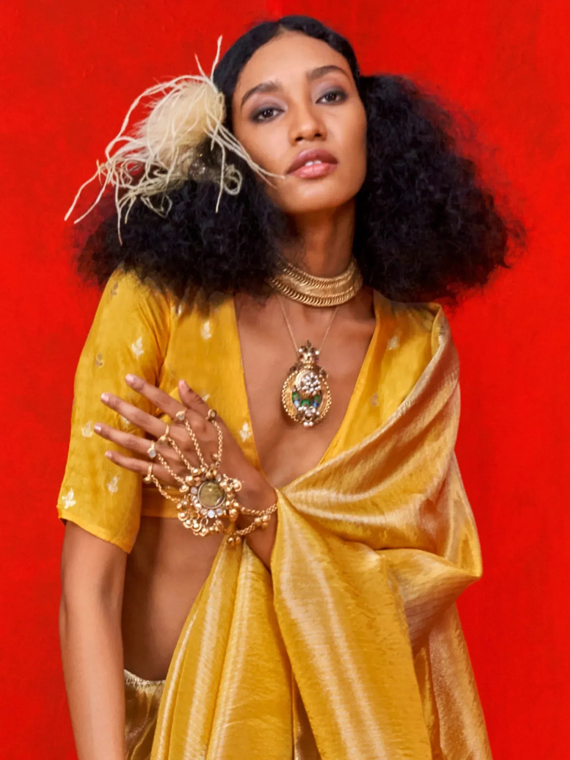 Woman in yellow ethnic saree with gold jewelry and feather hair accessory against red background