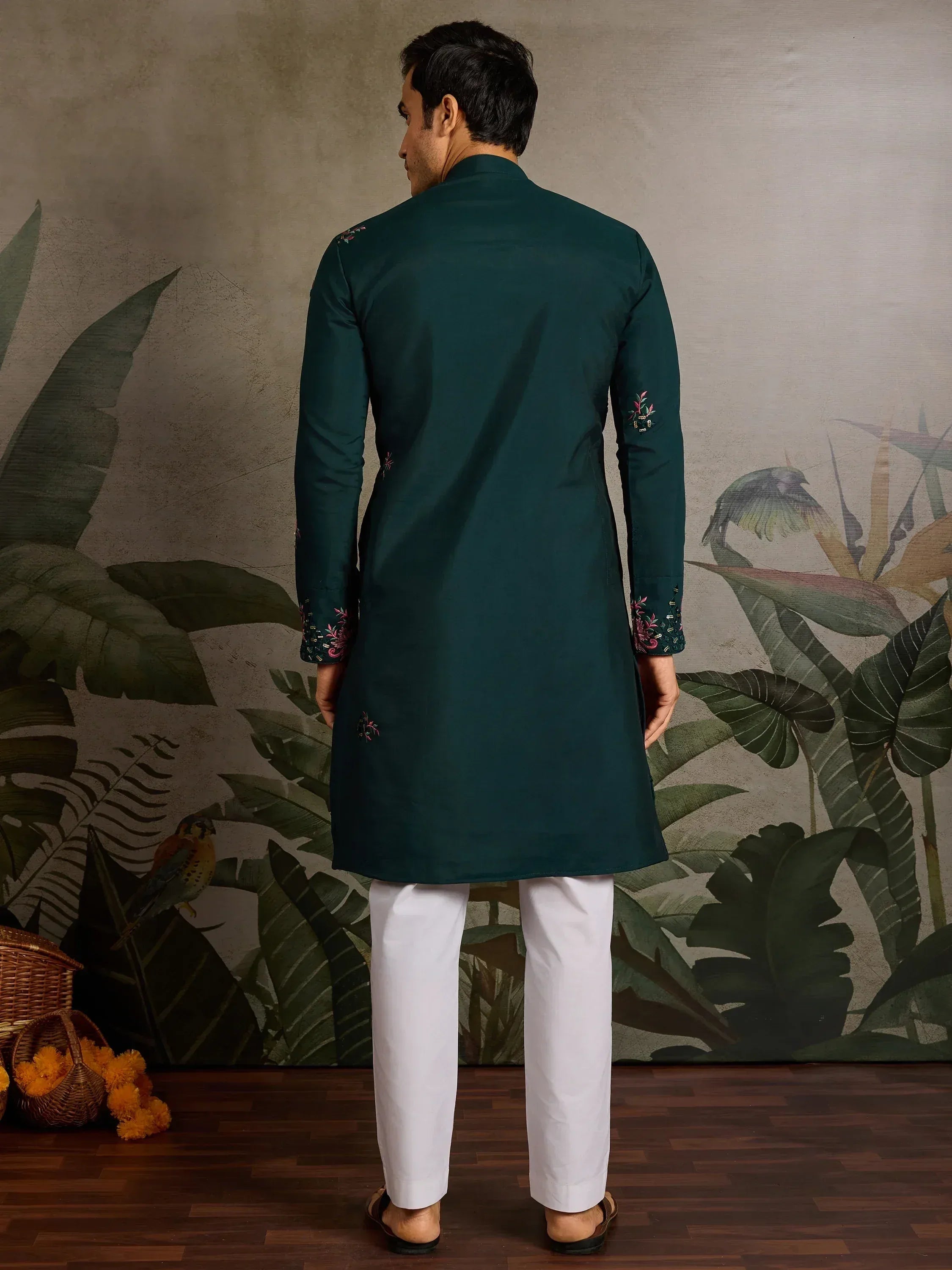 Man wearing dark green embroidered kurta with white pants, back view, tropical leaf backdrop
