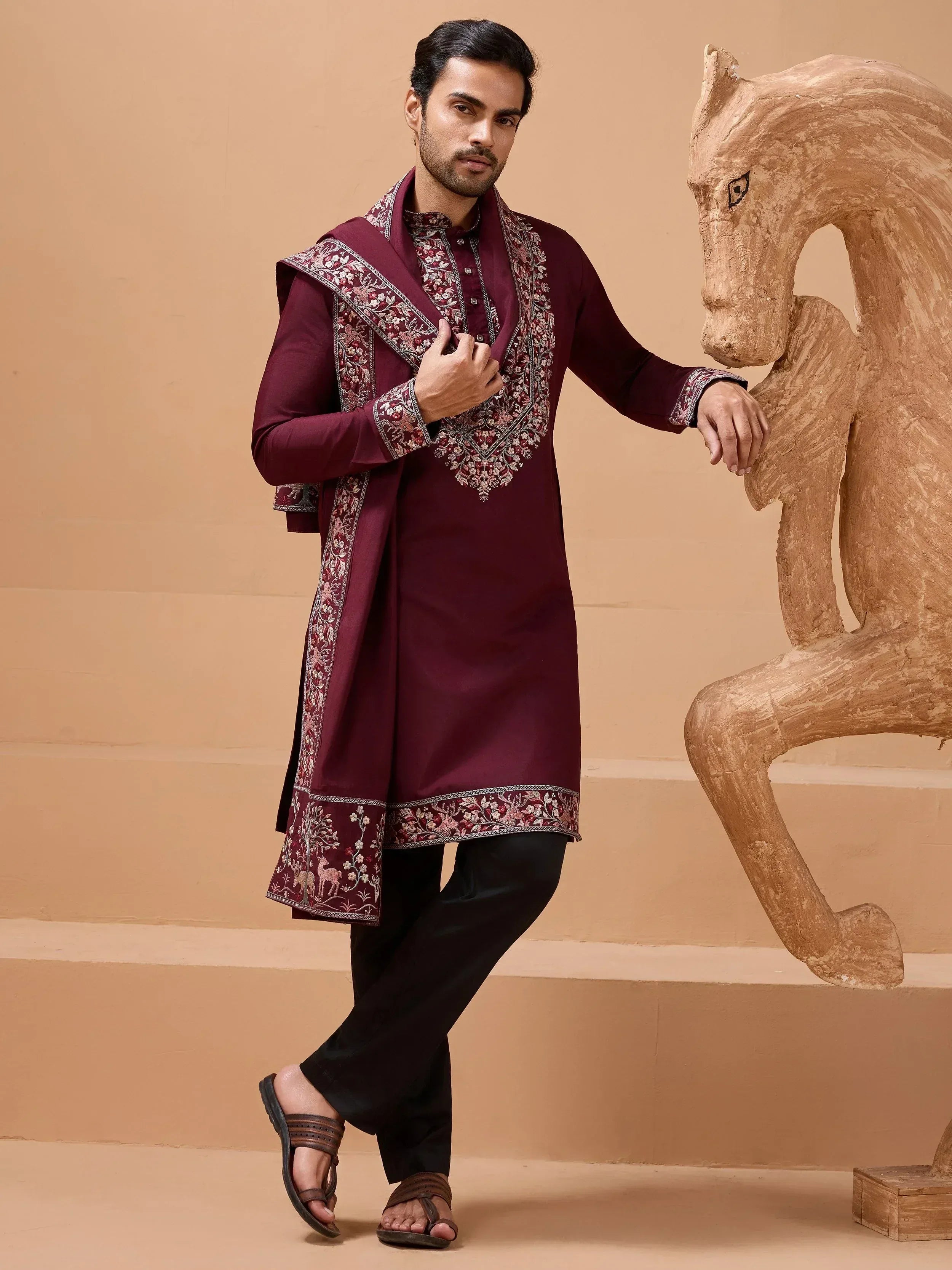 Man in maroon embroidered kurta with dupatta, black pants, and sandals, posing beside wooden horse sculpture