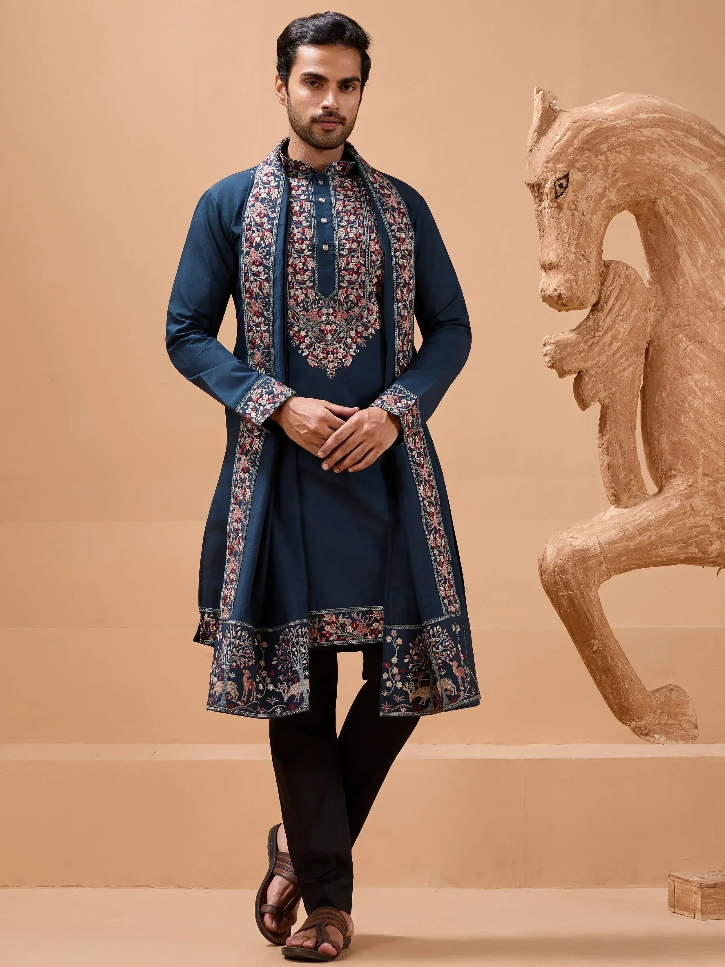 Man wearing navy blue embroidered kurta with floral and deer motifs, paired with black pants and sandals.