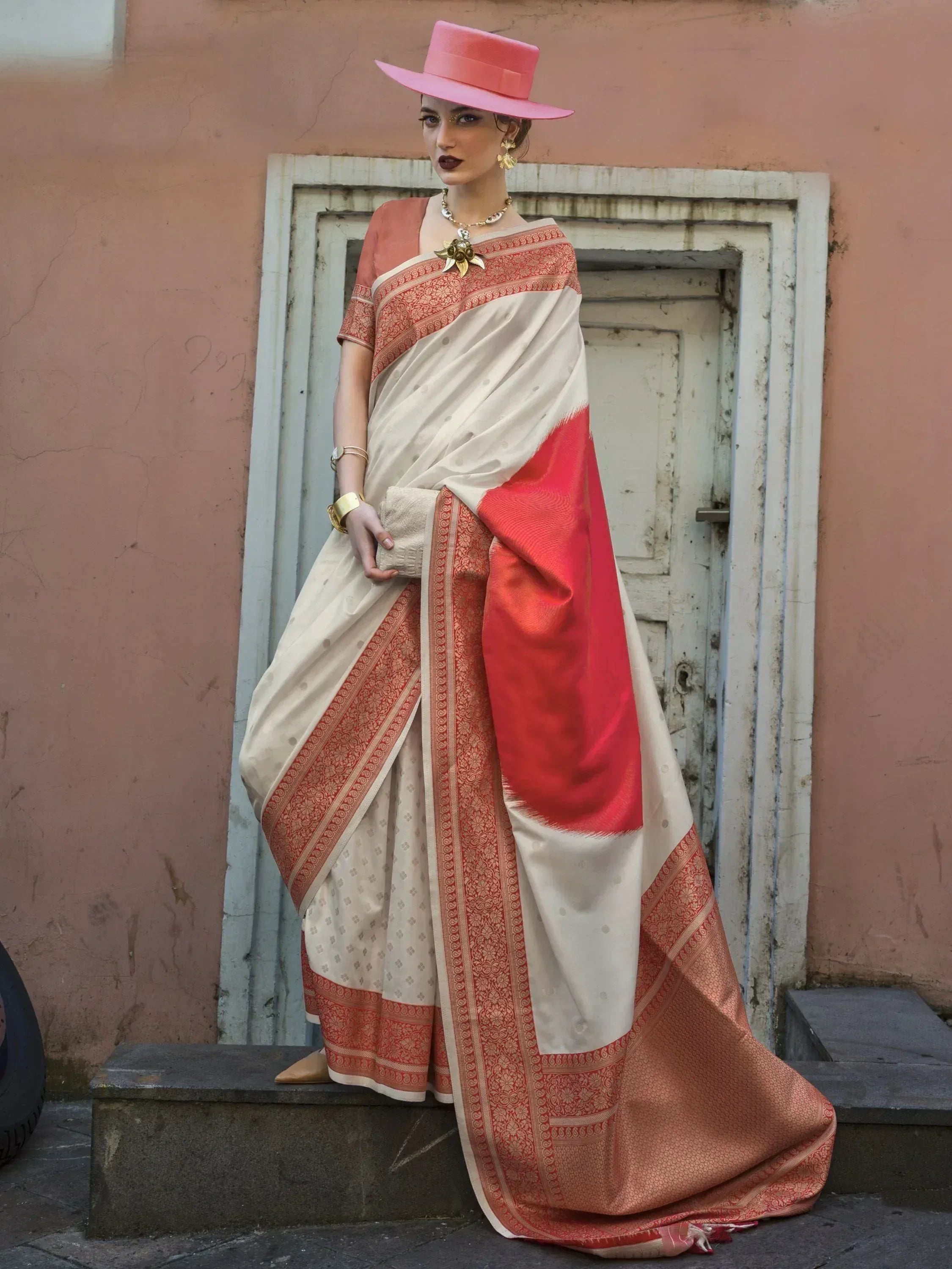 Woman in elegant cream and red saree with gold jewelry and pink hat, styled for ethnic fashion